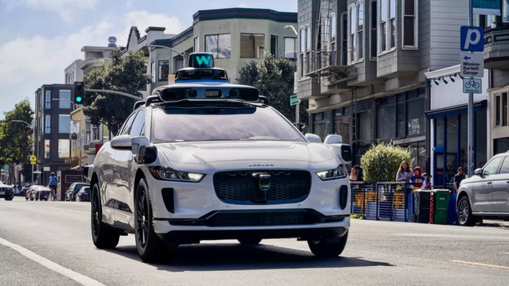 La compañía de robotaxis Waymo reconoce que a veces hay un filipino al volante Coche autónomo Jaguar blanco circulando por la ciudad bajo luz verde. Innovación en la movilidad urbana.