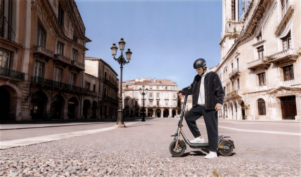 Xiaomi lanza patinetes Scooter 6, Watch 5, REDMI Buds 8 Pro y más gadgets Persona con casco monta patinete eléctrico en plaza adoquinada, rodeada de edificios históricos y cielo despejado.