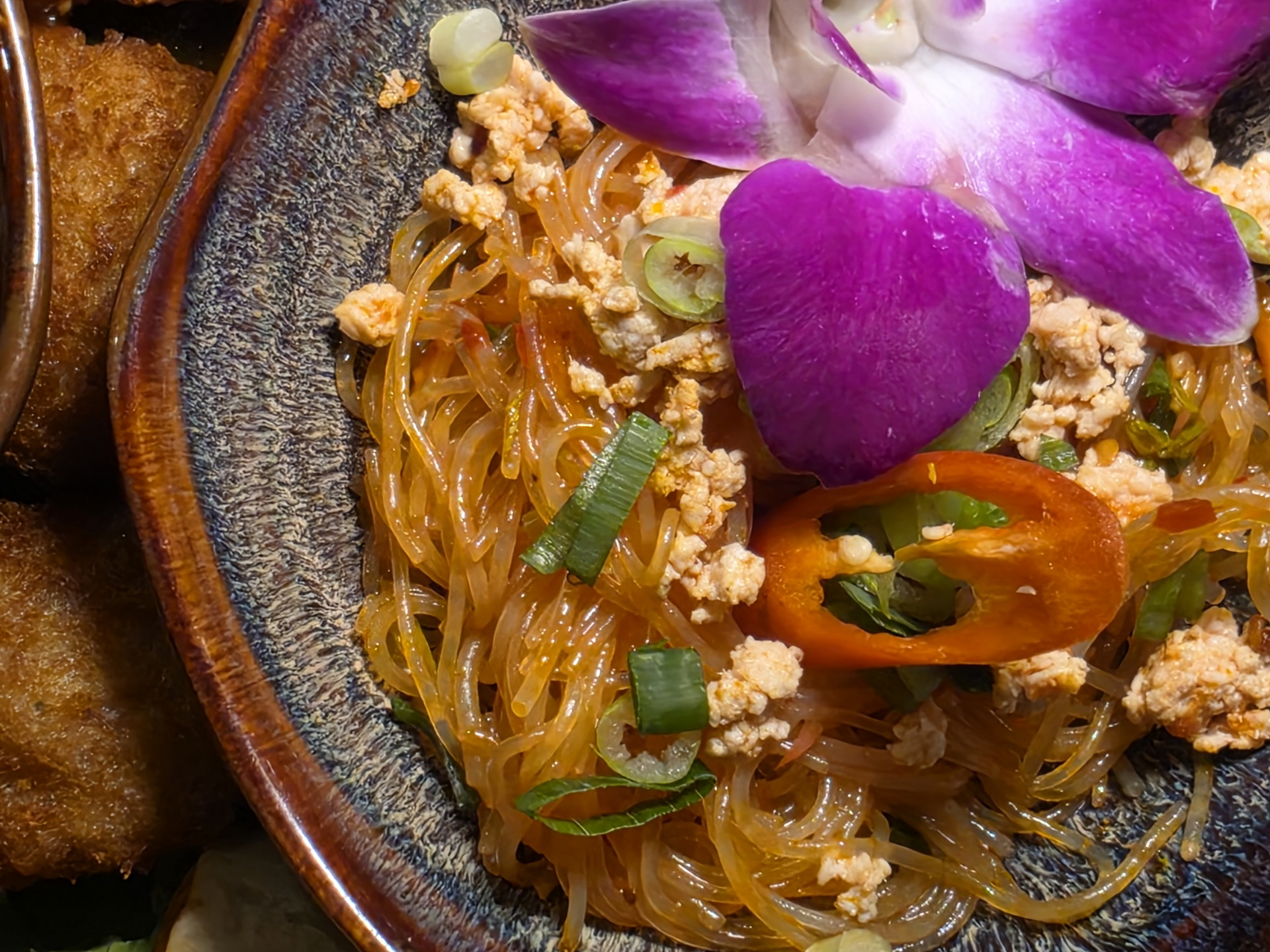 Plato de fideos de vidrio con carne picada, cebolla, chile rojo y una flor morada como decoración. Comida asiática.