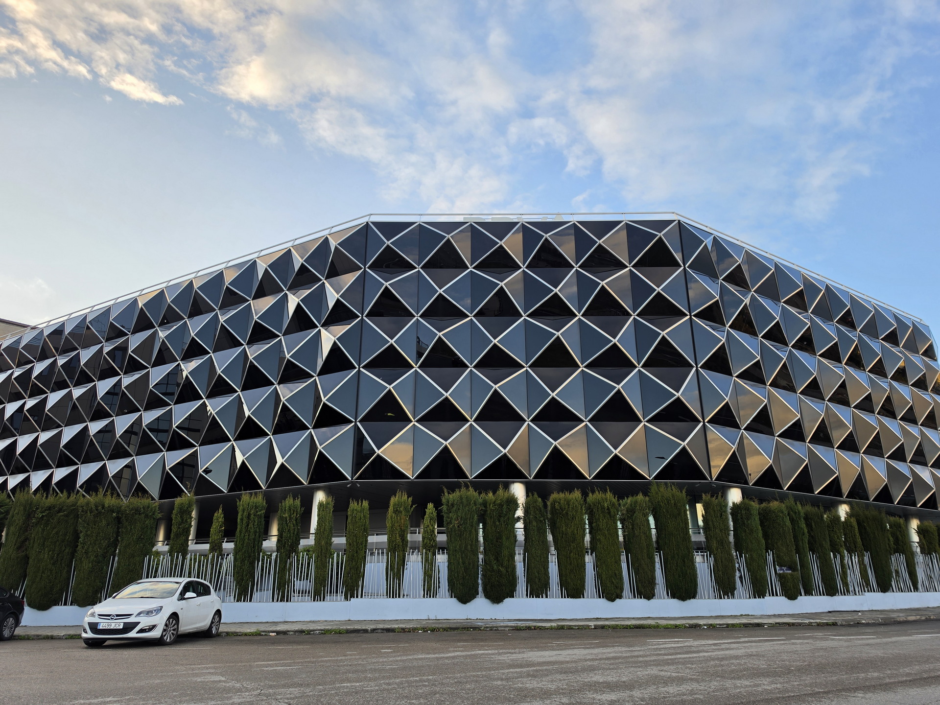 Edificio moderno con fachada geométrica y ventanas reflectantes al atardecer, cielo parcialmente nublado.