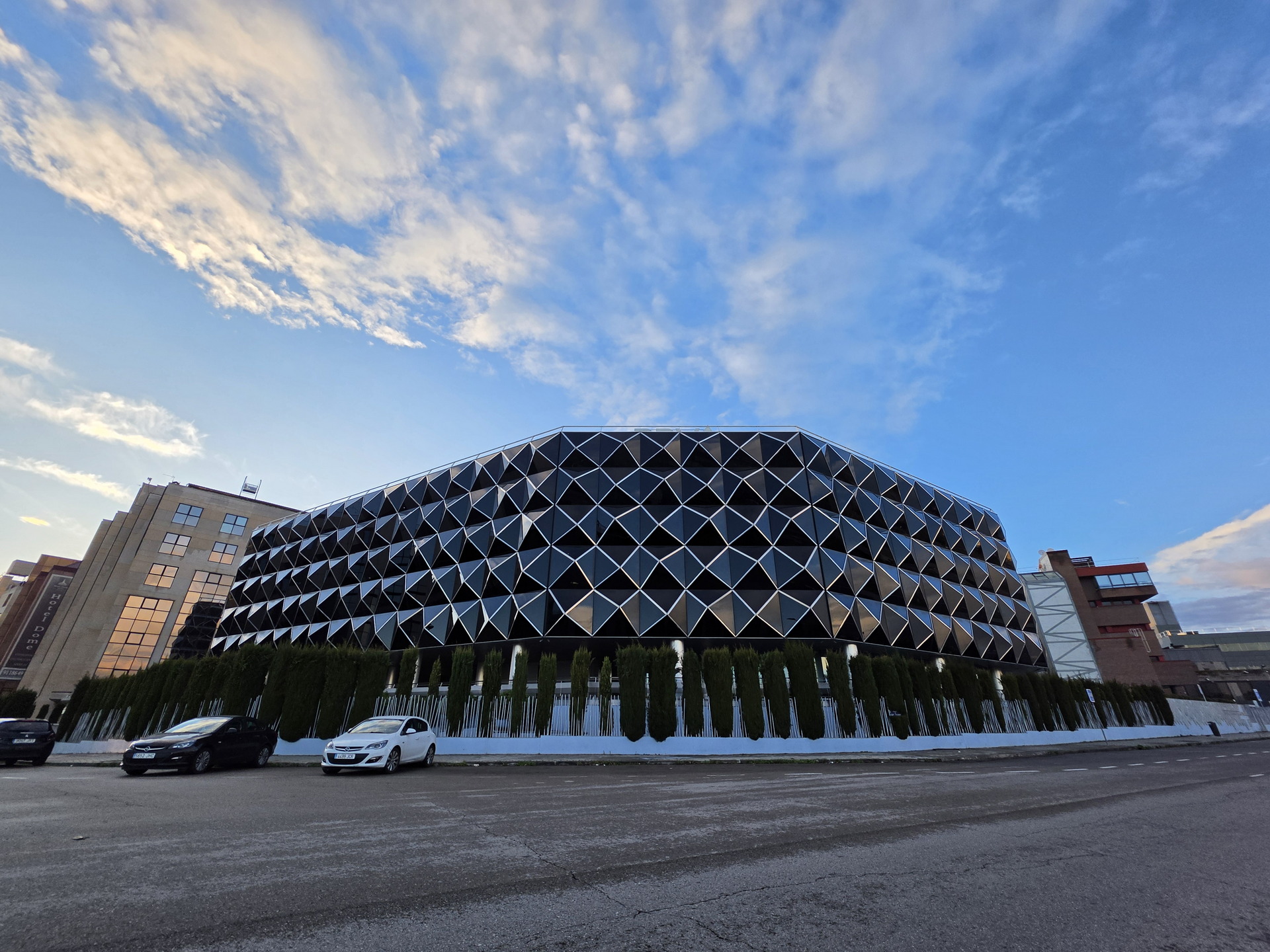 Edificio moderno con fachada geométrica bajo cielo azul y coches estacionados enfrente, al atardecer.