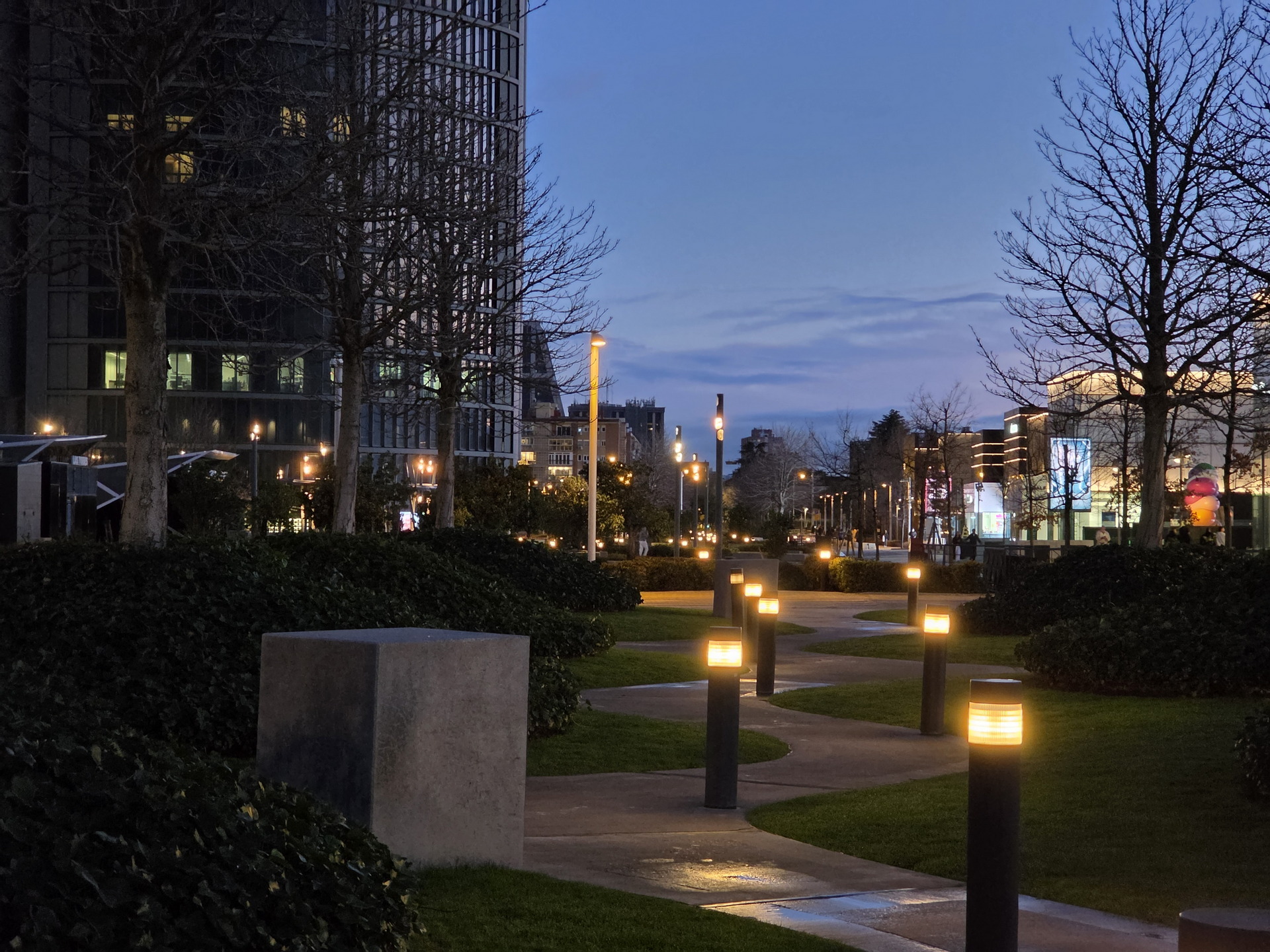 Parque urbano al anochecer con luces encendidas y caminos iluminados cerca de edificios modernos.