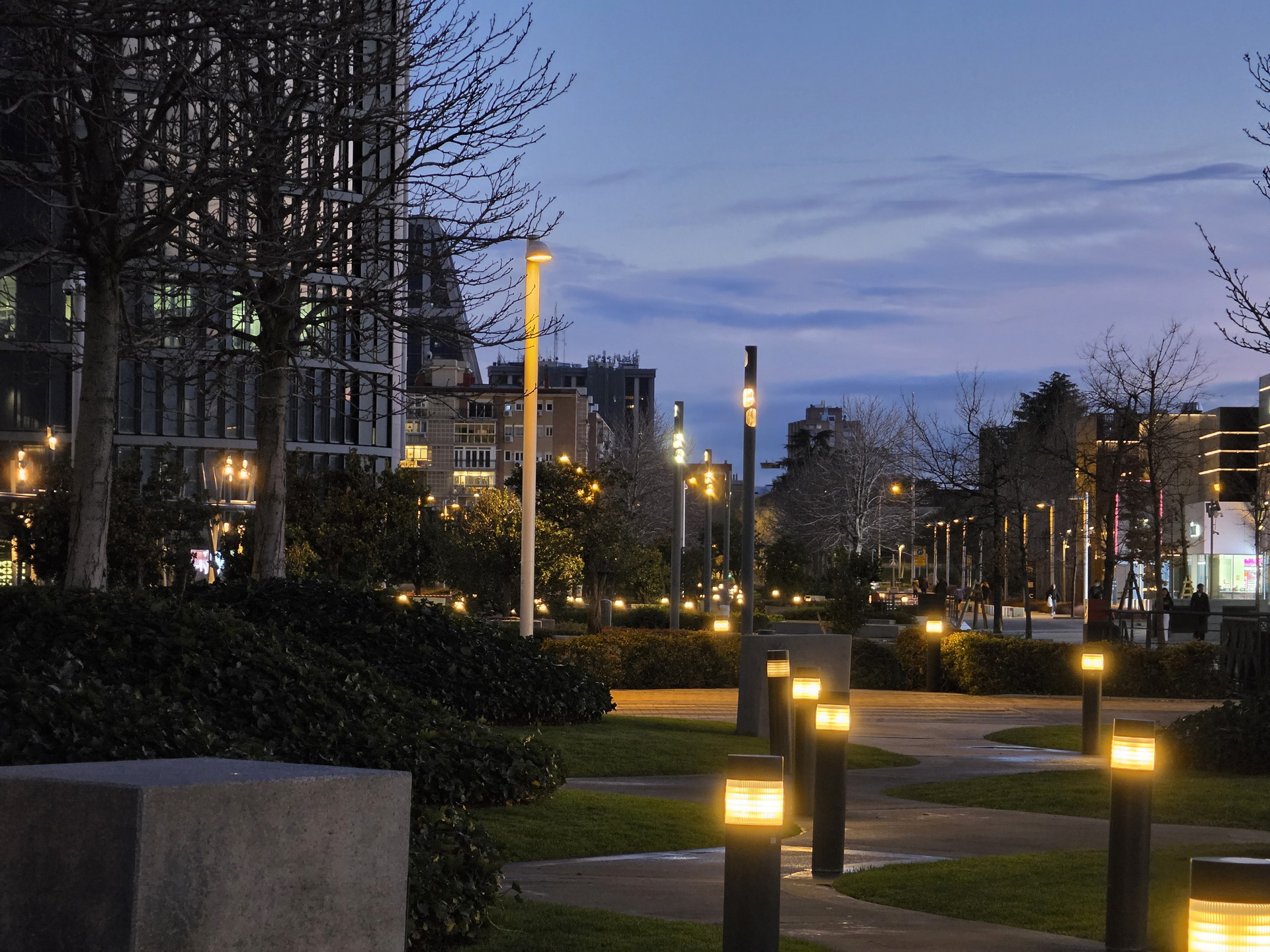 Parque urbano iluminado al anochecer con bancos, árboles y edificios al fondo, creando una atmósfera tranquila.