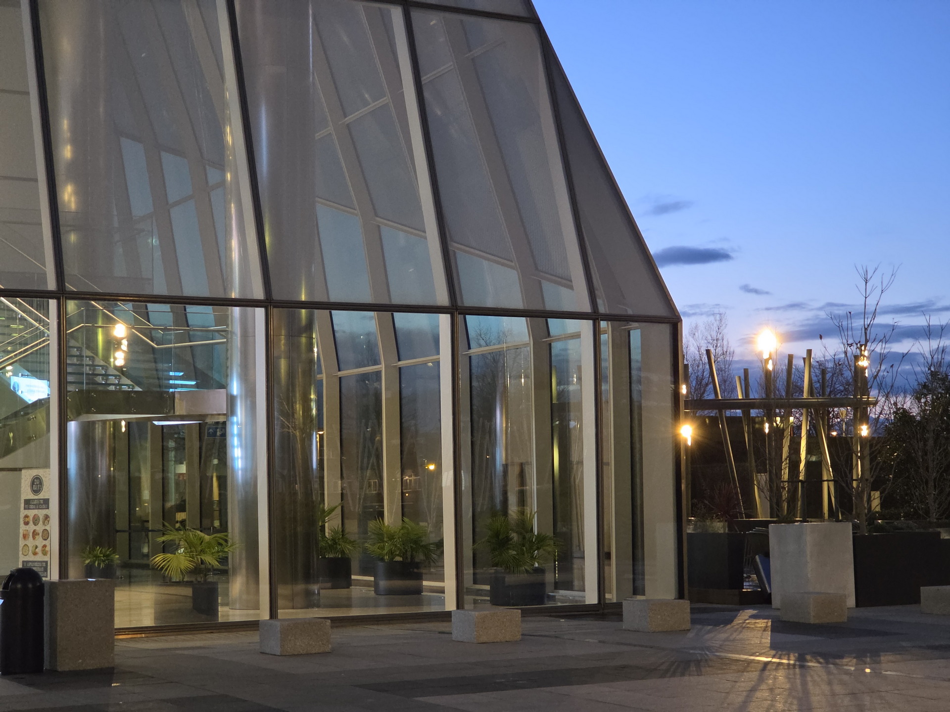 Entrada moderna de edificio con fachada de vidrio al anochecer, luces encendidas y plantas decorativas.