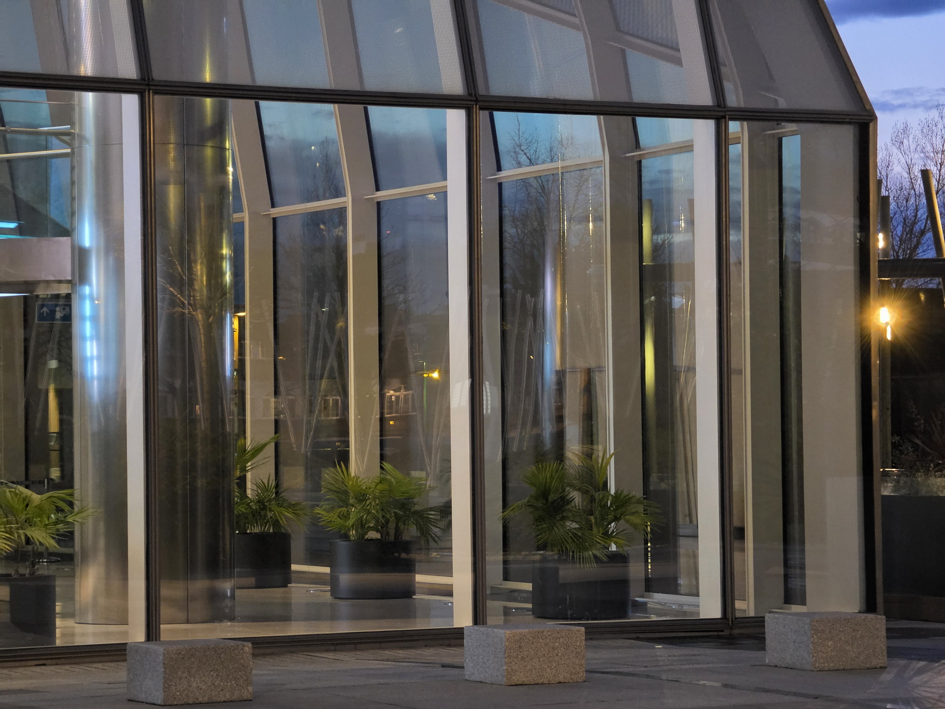 Edificio moderno de cristal con plantas en el interior, reflejando luces durante el atardecer.