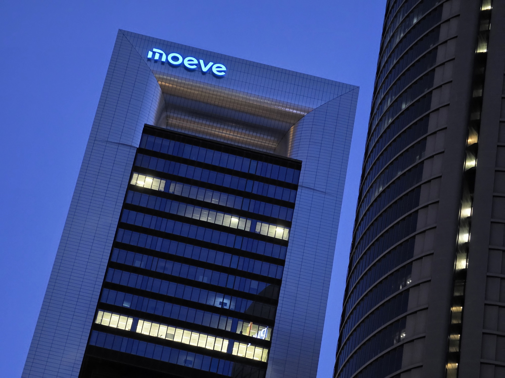 Rascacielos iluminado al anochecer con el logo de moeve en la cima, arquitectura moderna en contraste con el cielo azul.