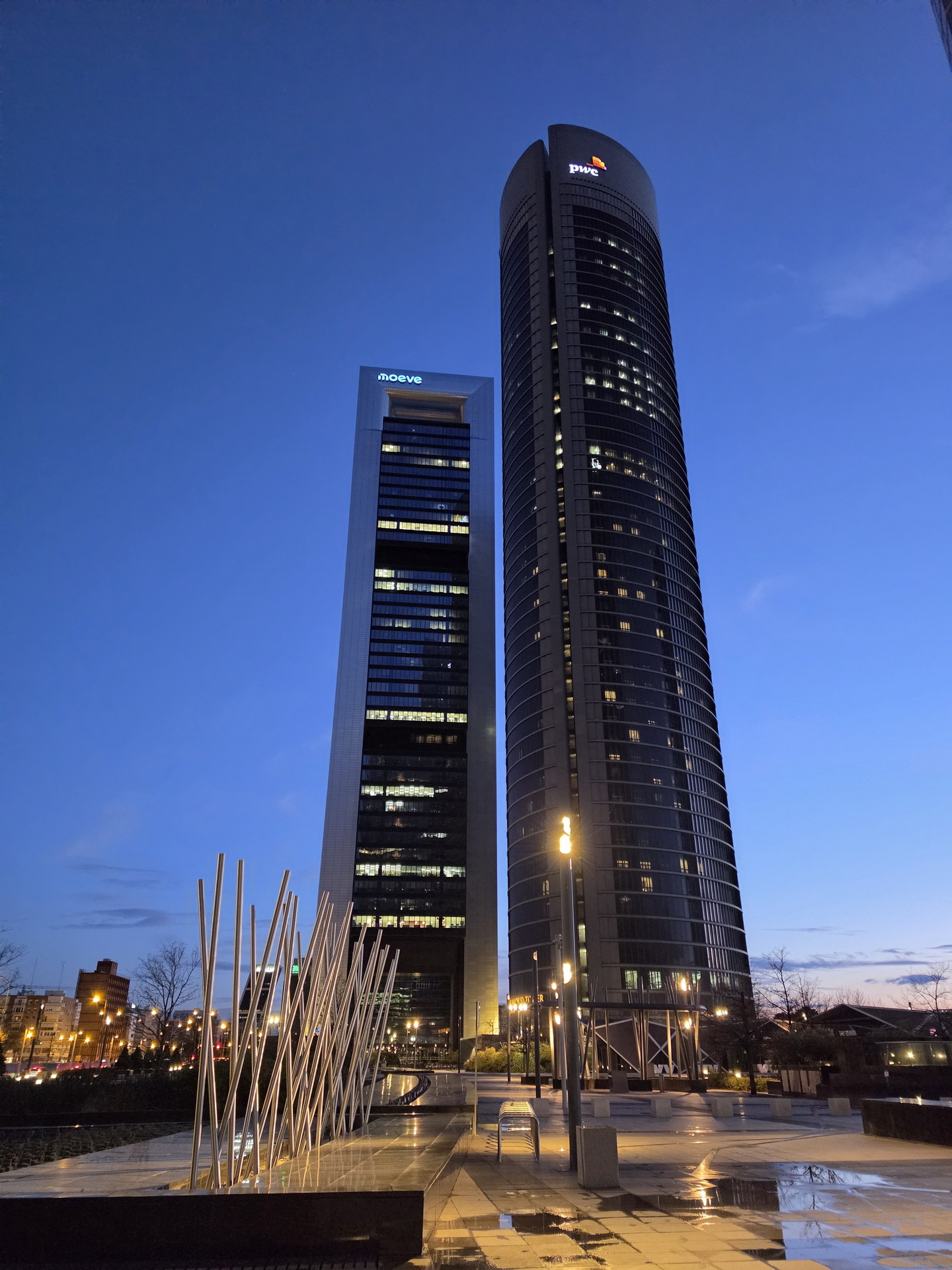 Torres modernas al atardecer en ciudad iluminada, cielo azul y escultura en primer plano.