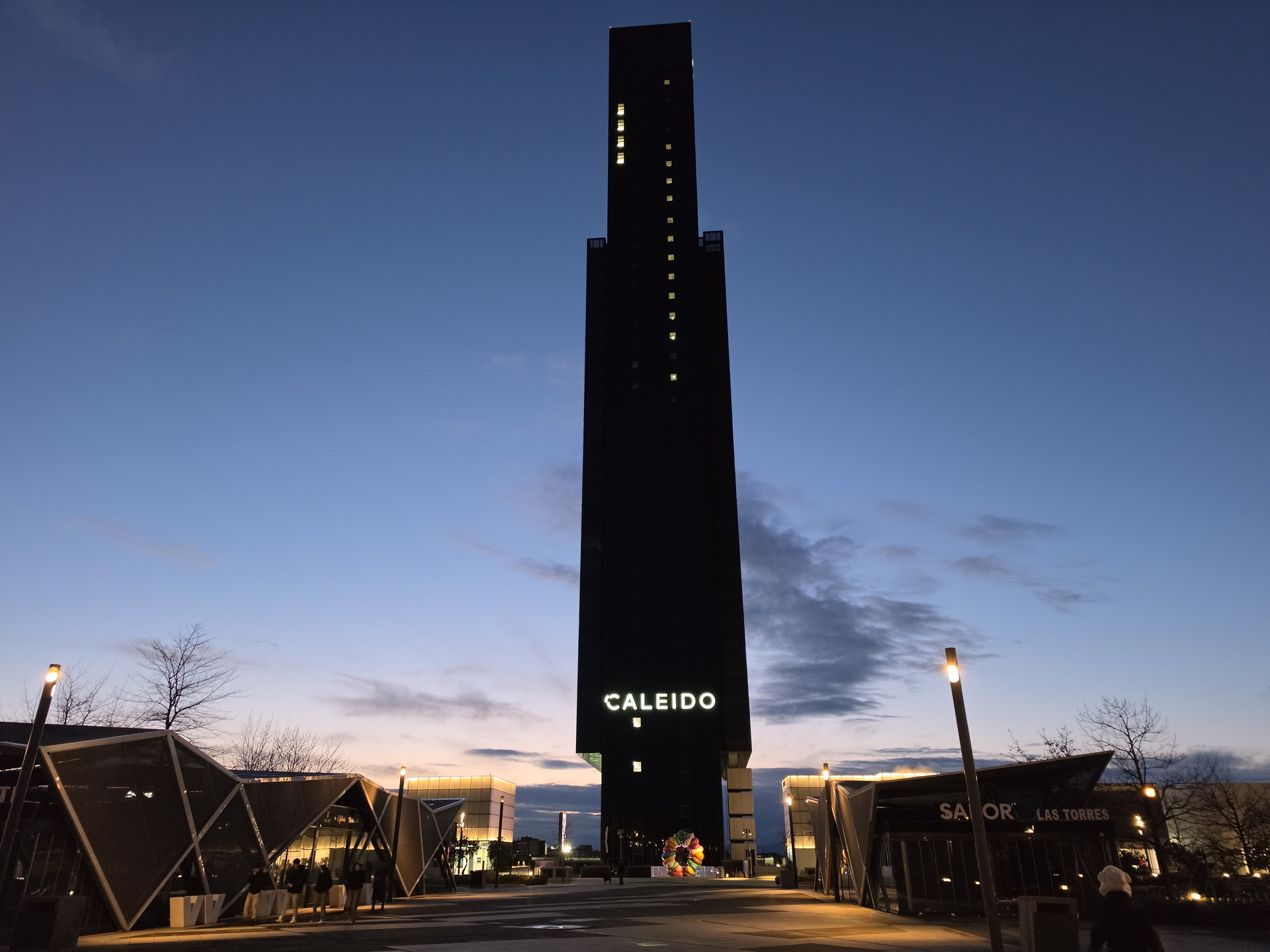 Edificio Caleido iluminado al atardecer, con cielo azul y luces encendidas, Madrid.