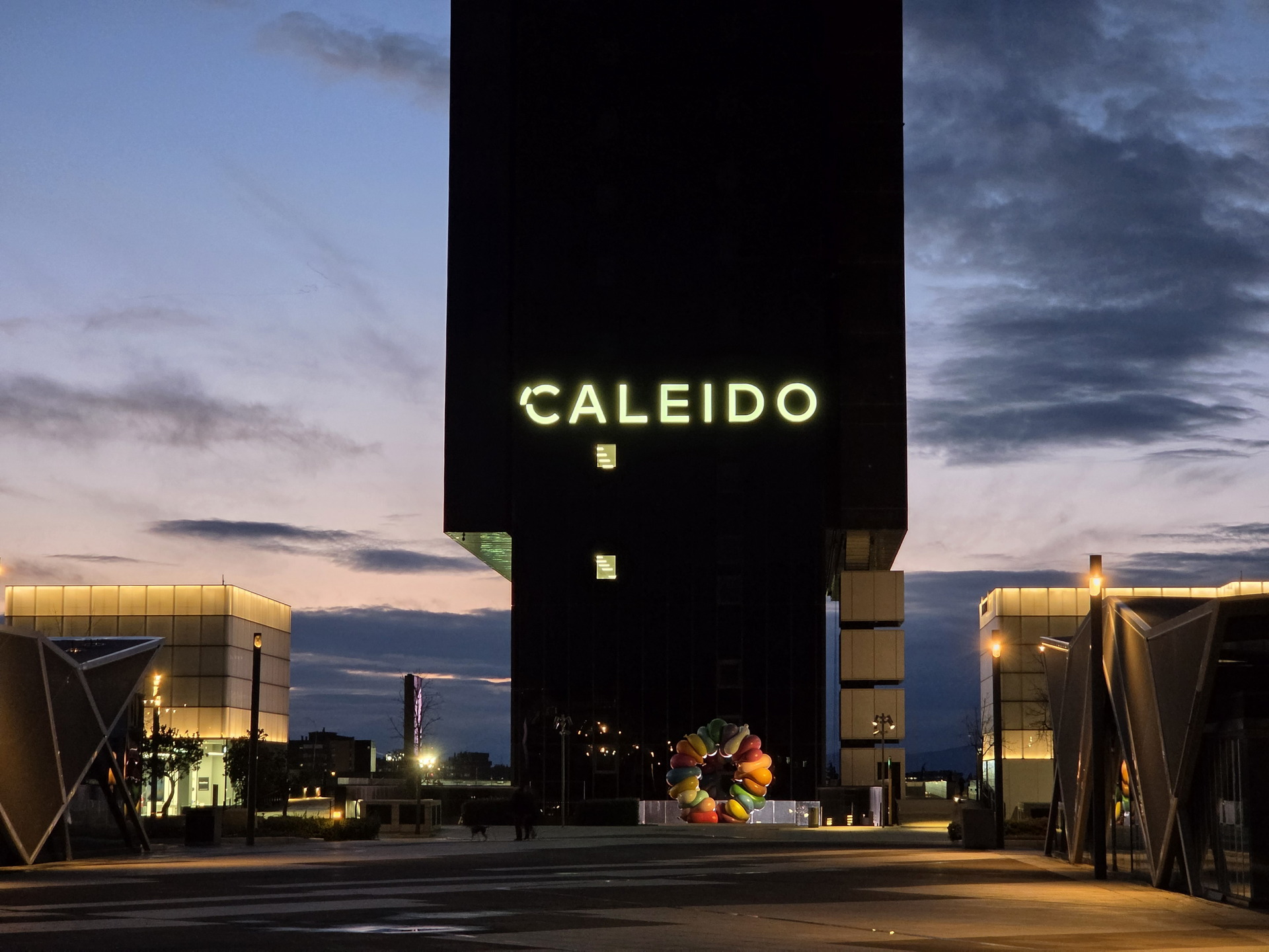 Edificio Caleido iluminado al atardecer en Madrid, con cielo nublado y estructuras modernas alrededor.