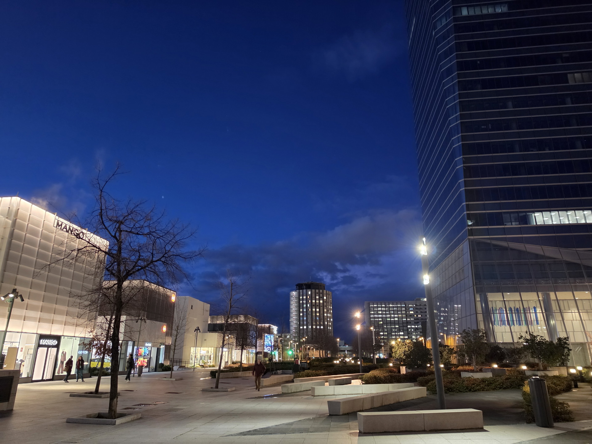 Vista nocturna de modernos edificios iluminados y amplias calles peatonales en una ciudad, con cielo azul profundo.