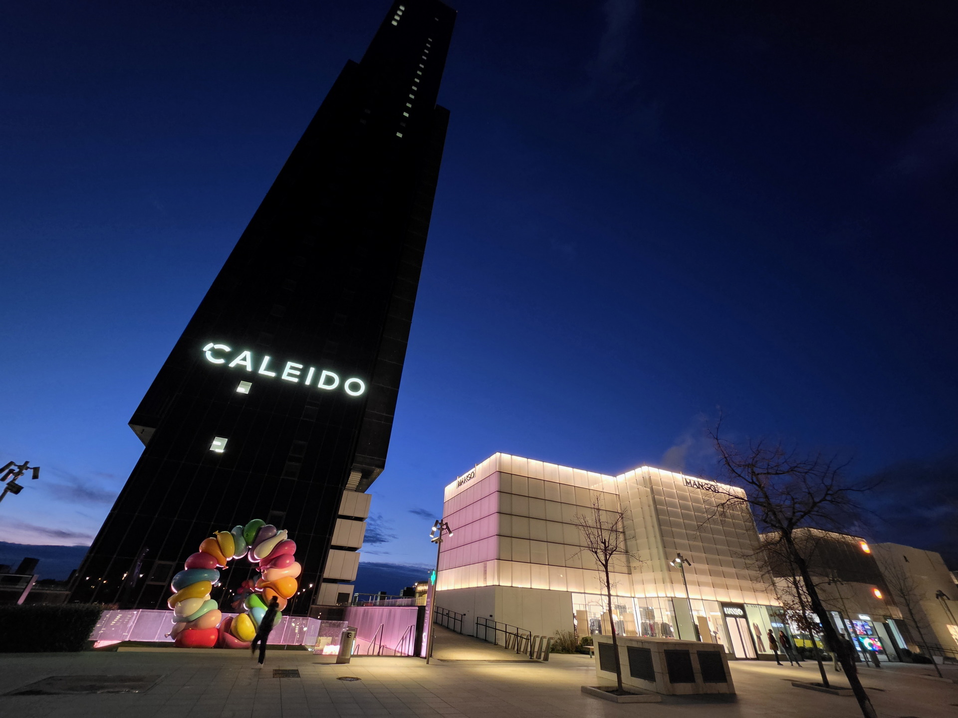 Torre Caleido Madrid: Edificio iluminado al anochecer junto a escultura de colores vivos.