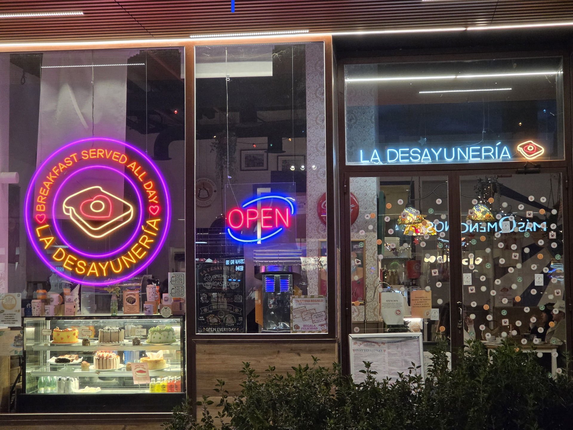 Fachada de cafetería La Desayunería con letreros de neón y vitrinas de postres. Abierto todo el día.