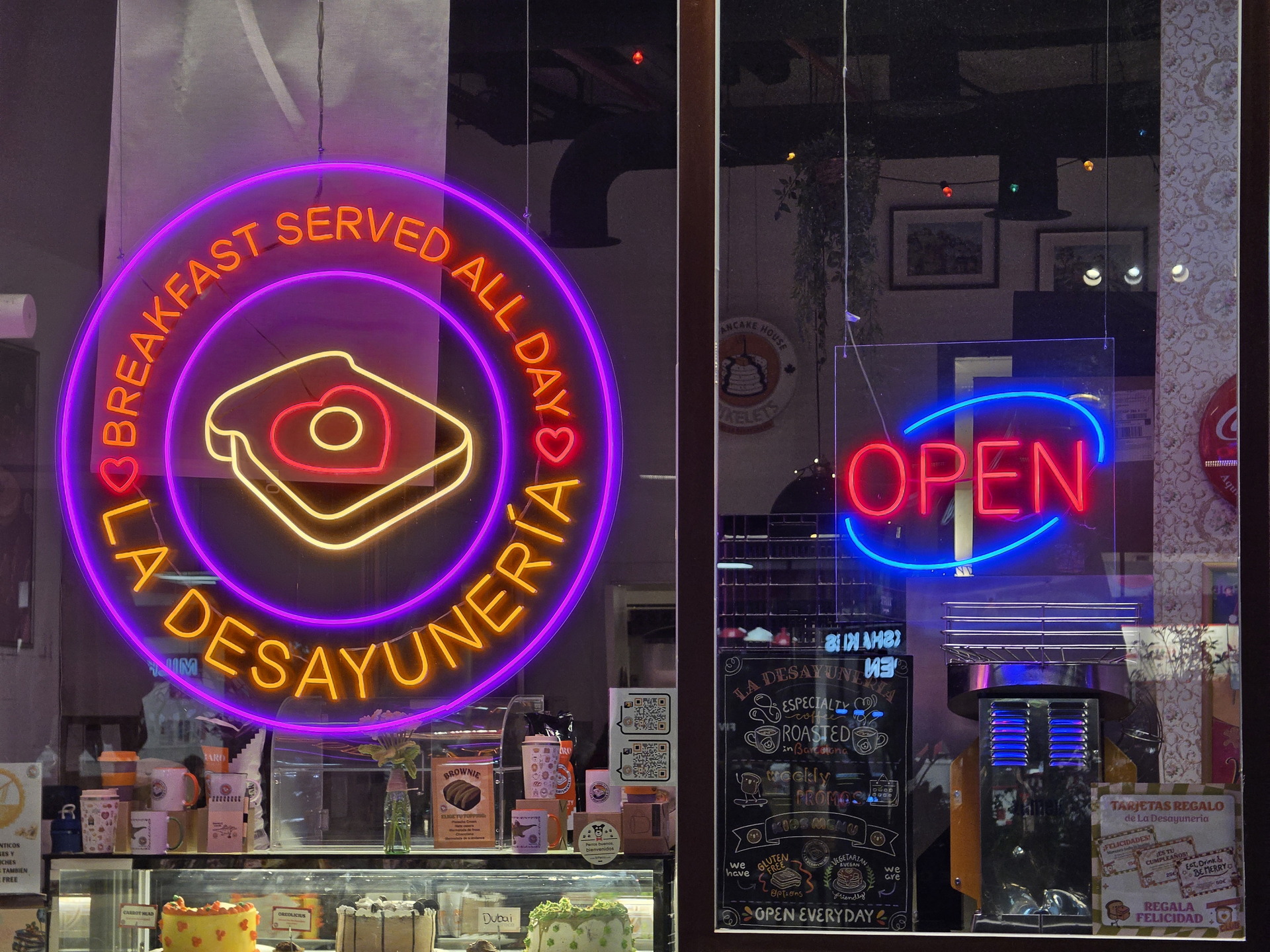 Letrero de neón La Desayunería y Open en una ventana, desayuno servido todo el día. Decoración interior visible.