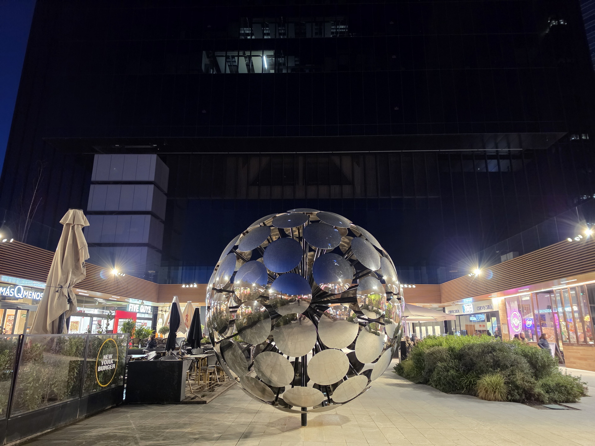 Escultura esférica metálica refleja luces nocturnas en plaza urbana con tiendas y restaurantes.