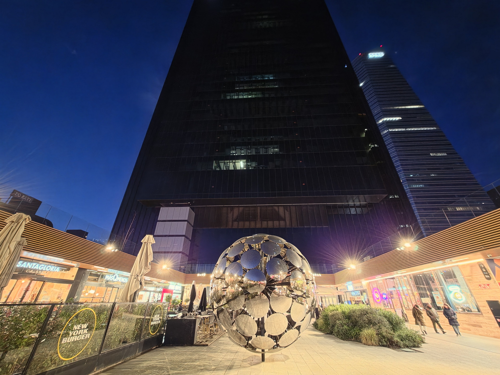 Plaza urbana al anochecer con escultura esférica y rascacielos iluminados en el fondo.