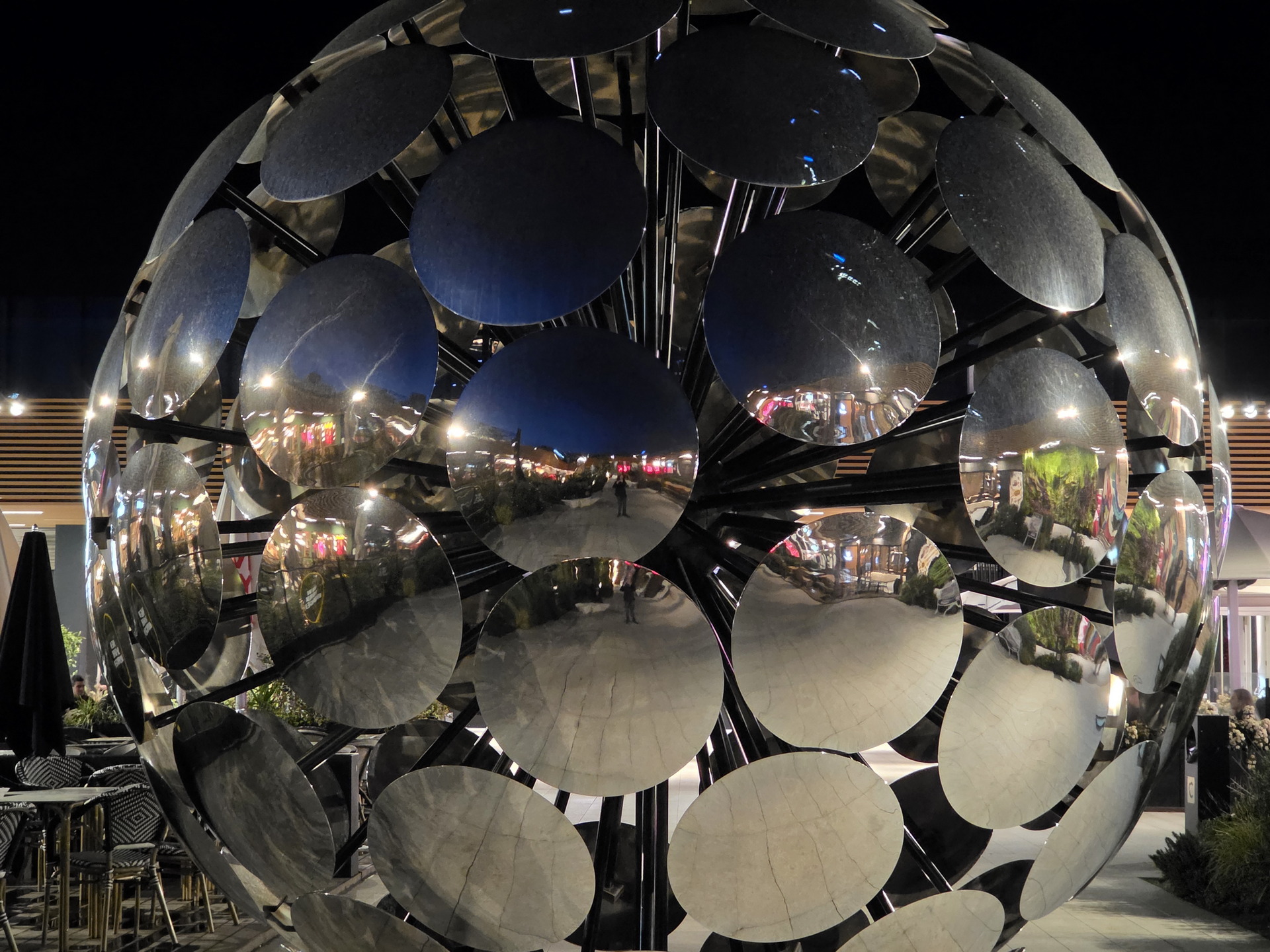 Escultura esférica de espejos reflejando luces de ciudad por la noche. Reflejos multicolores y arquitectura moderna.