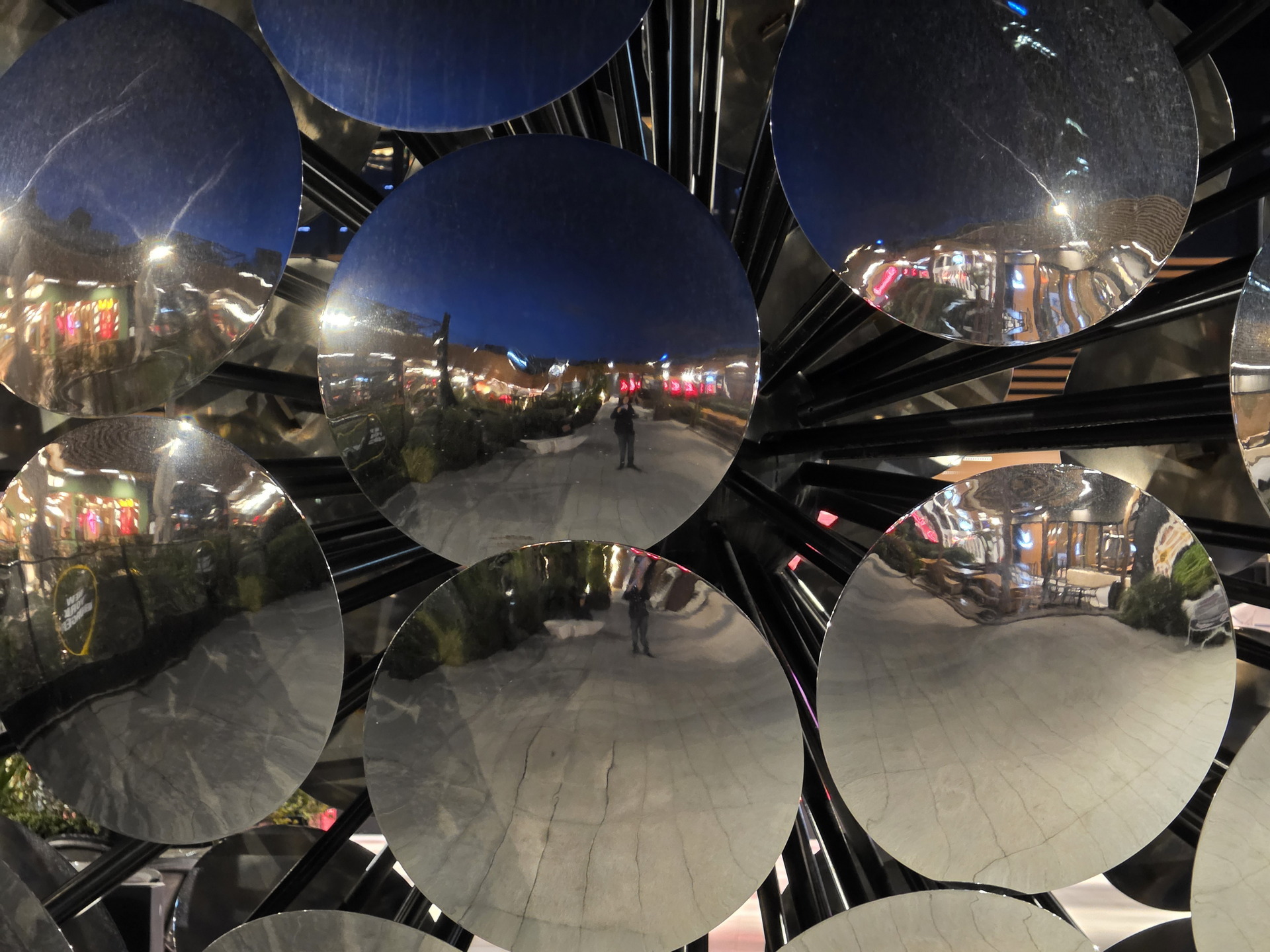 Reflejo urbano nocturno en discos metálicos, luces de ciudad y peatones creando un collage visual.