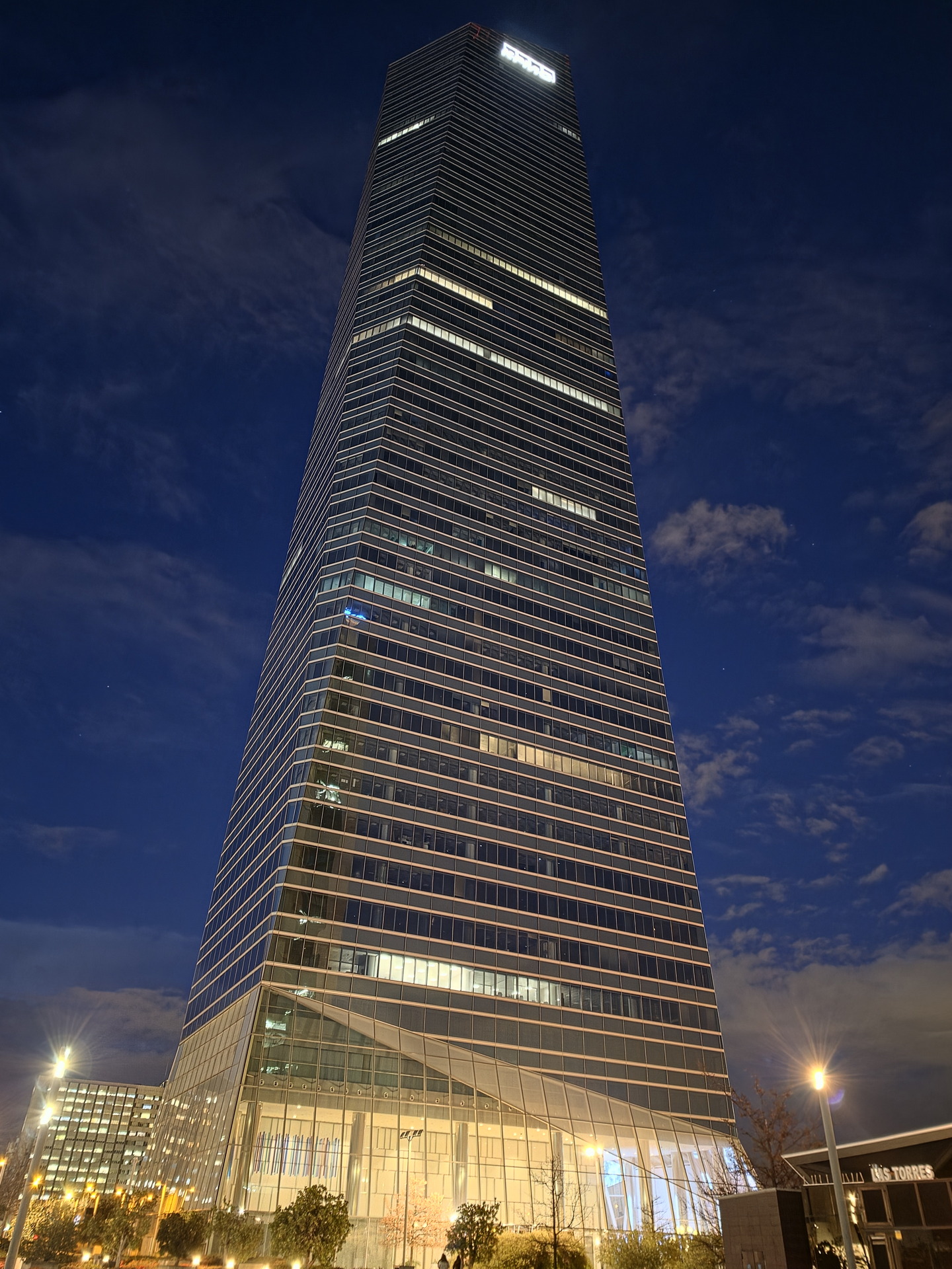 Rascacielos iluminado al anochecer con cielo despejado y algunas nubes, vista desde la base hacia la cima del edificio.