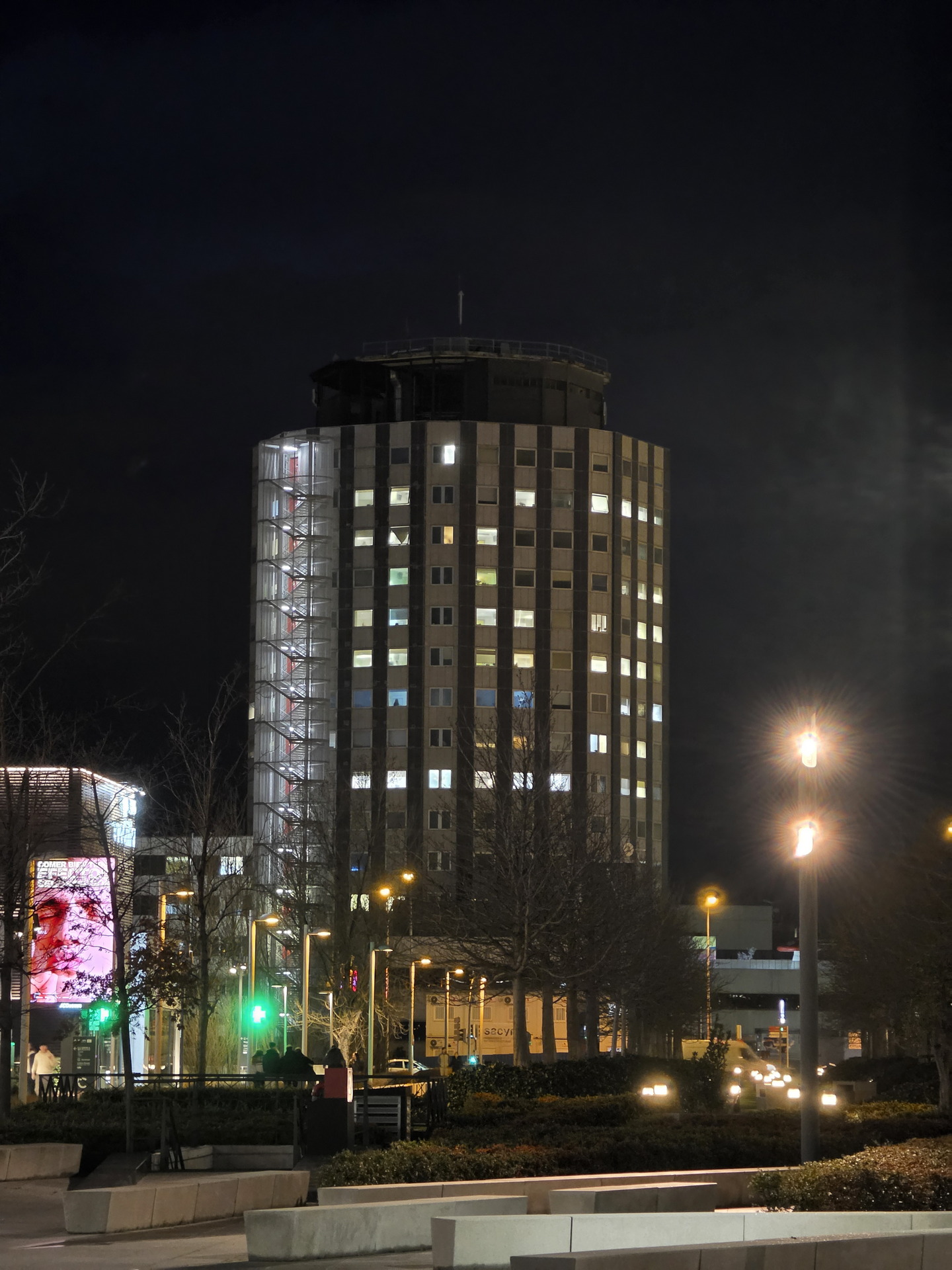 Edificio iluminado de noche en un área urbana con luces de calles y árboles en una ciudad moderna.