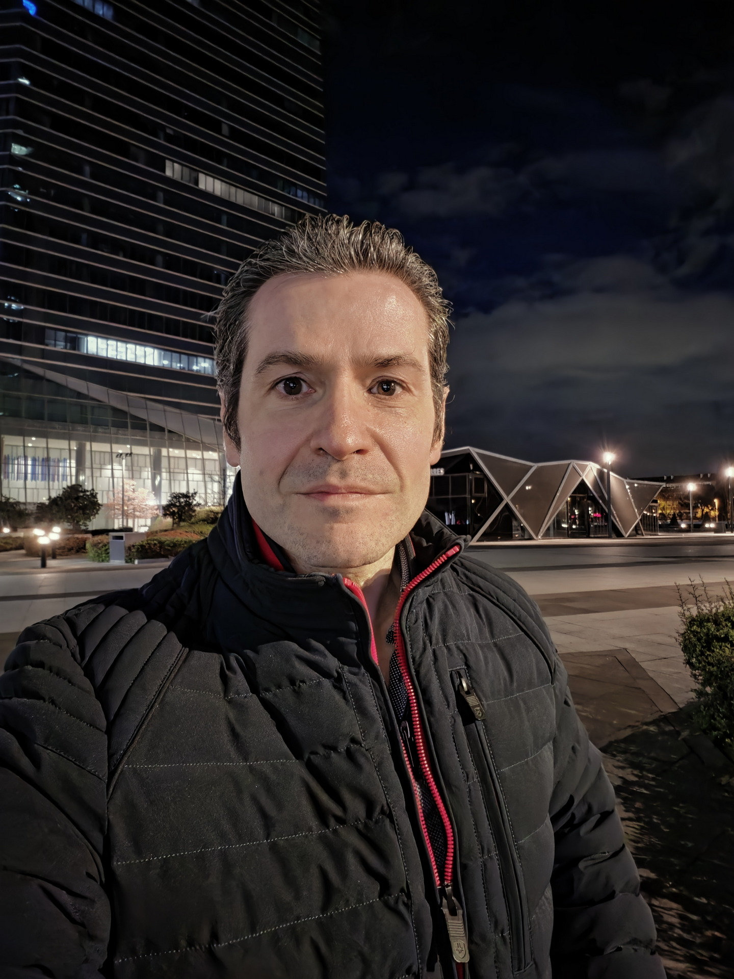Hombre con chaqueta negra en selfie nocturno frente a moderno edificio iluminado, cielo oscuro de fondo.