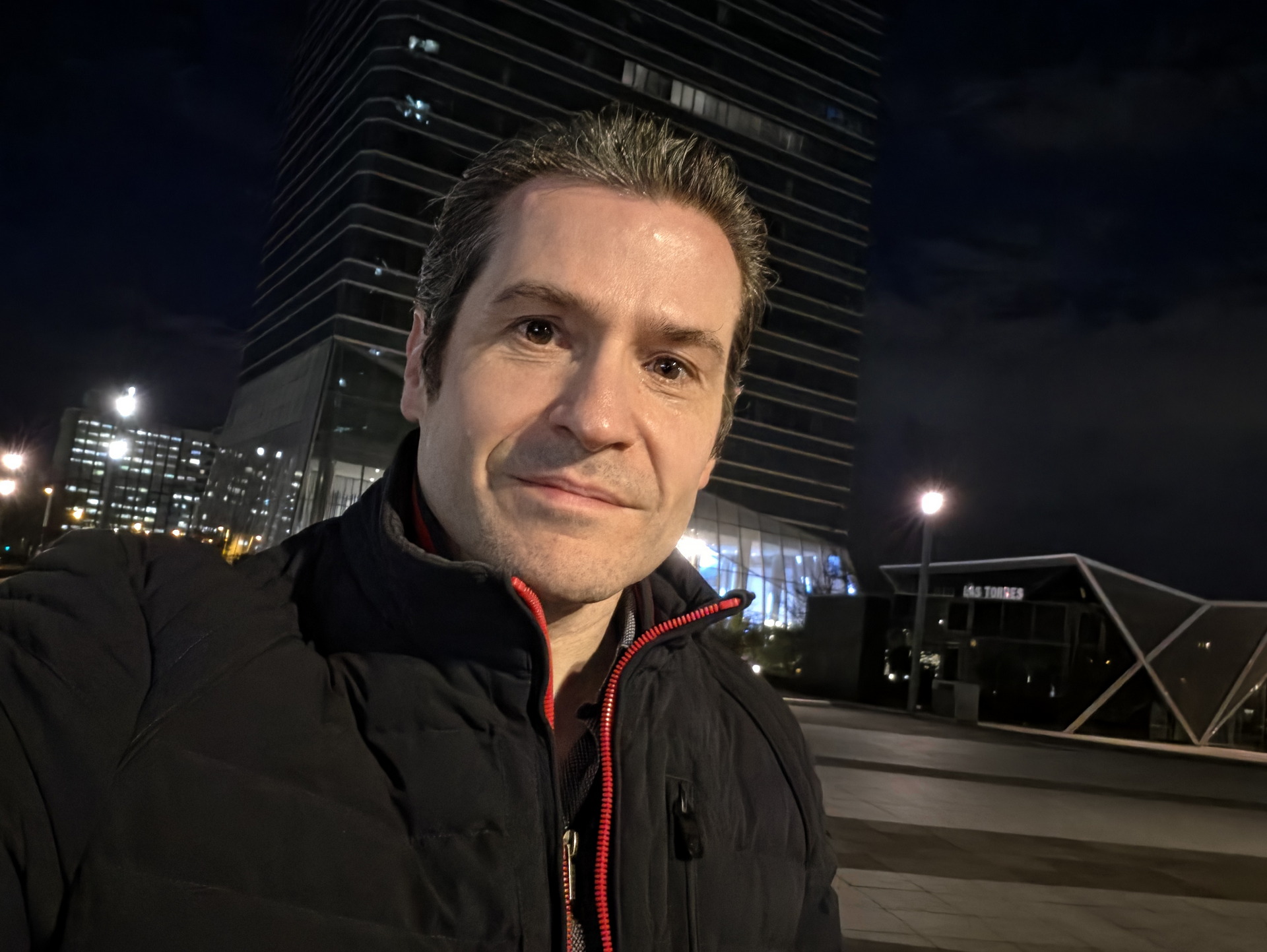 Hombre con chaqueta negra en selfie nocturno frente a edificio moderno iluminado.