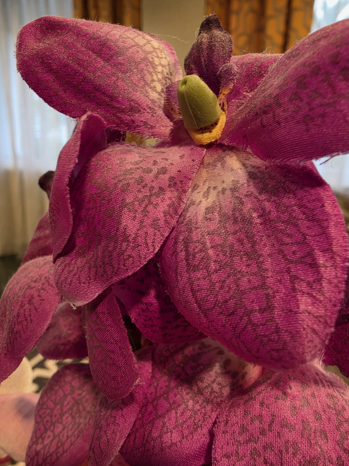 Flor artificial violeta con textura detallada y fondo difuminado en interior iluminado por luz natural.