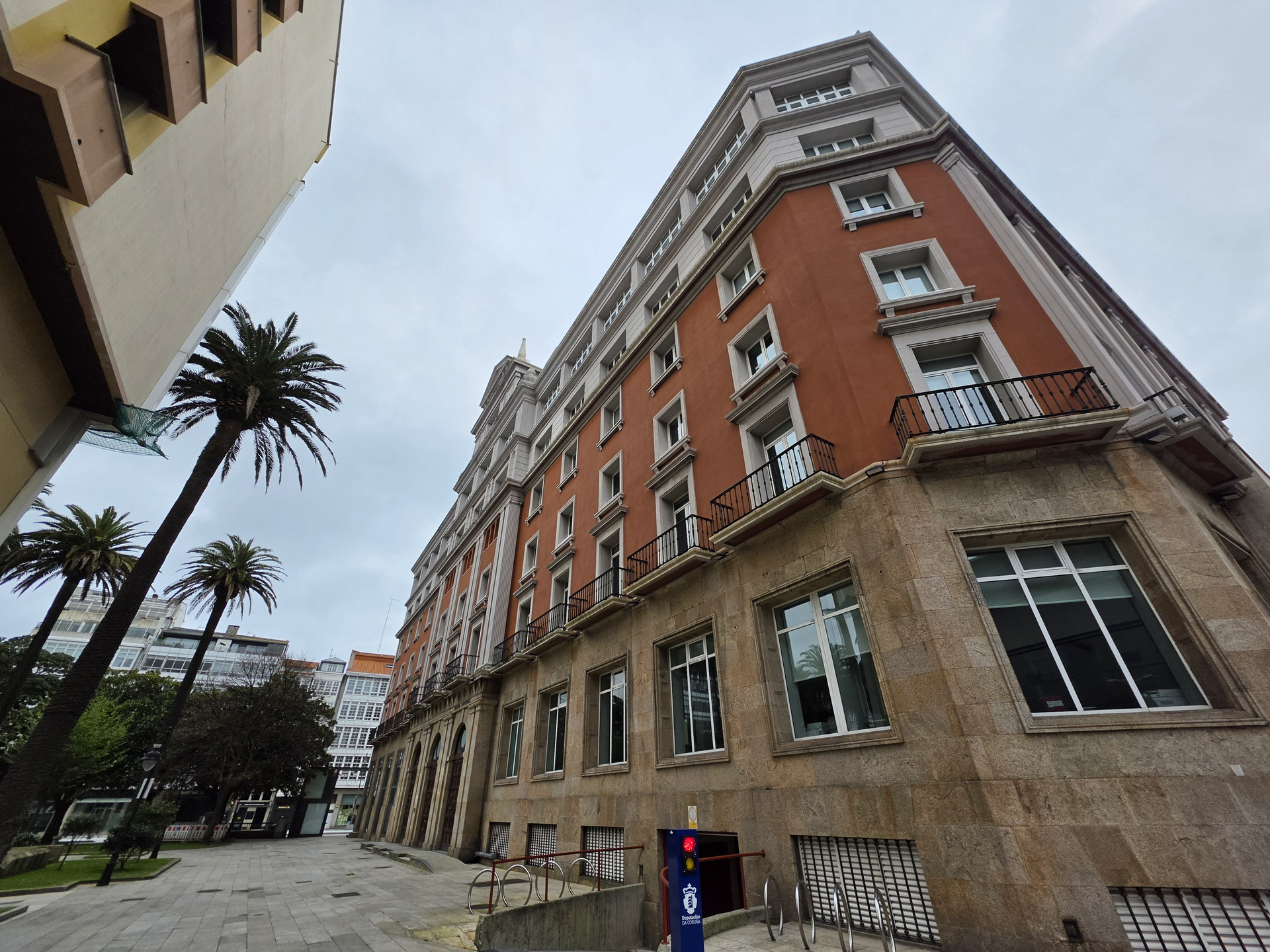 Edificio de fachada roja y ventanas blancas, rodeado de palmeras, bajo un cielo nublado en un área urbana.