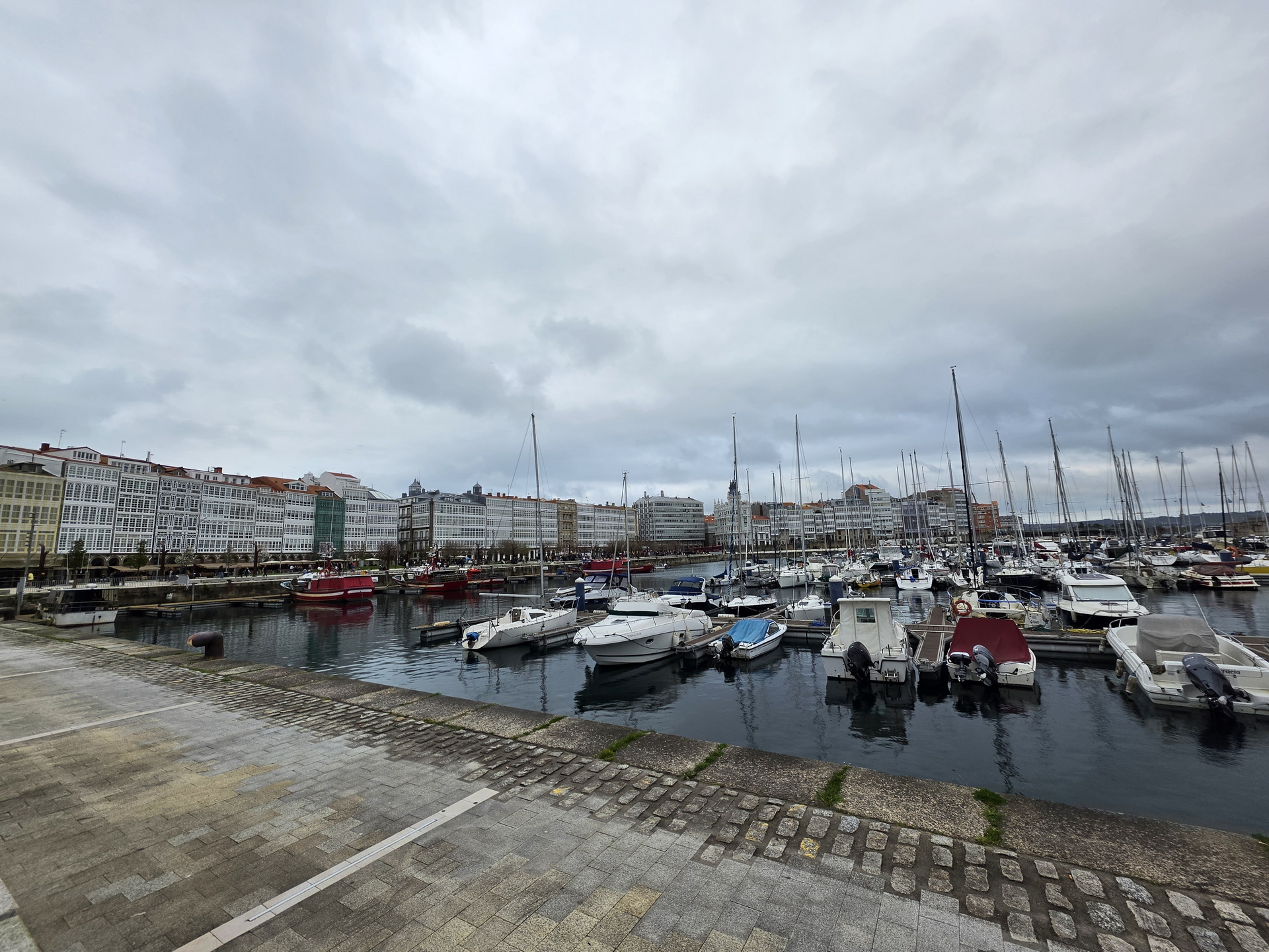 Puerto deportivo con barcos y edificios acristalados al fondo en día nublado, en la ciudad de La Coruña.