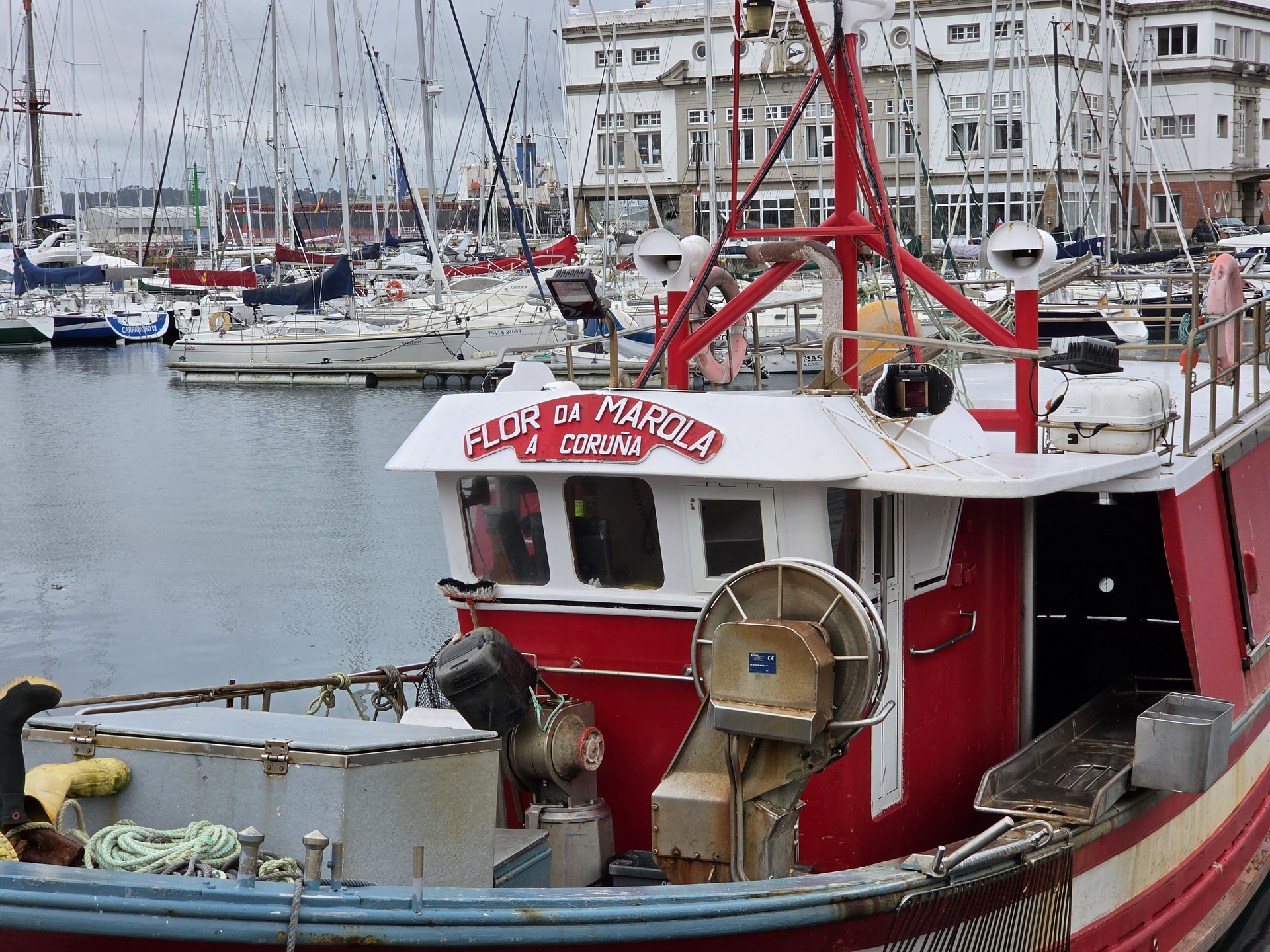 Barco pesquero rojo Flor da Marola en puerto de A Coruña, rodeado de veleros y edificios costeros.