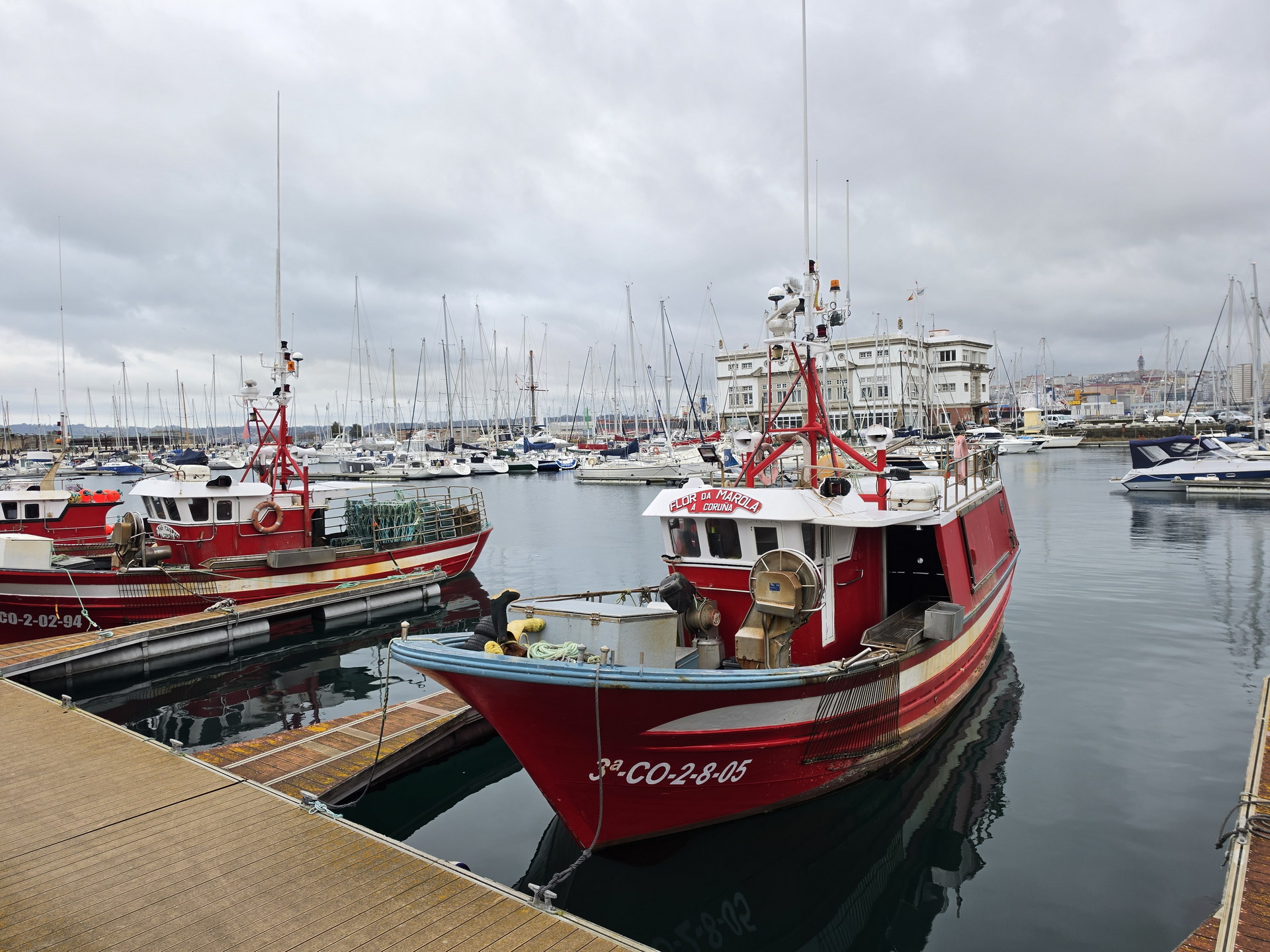 Barcos de pesca rojos atracados en un puerto con yates al fondo en un día nublado.
