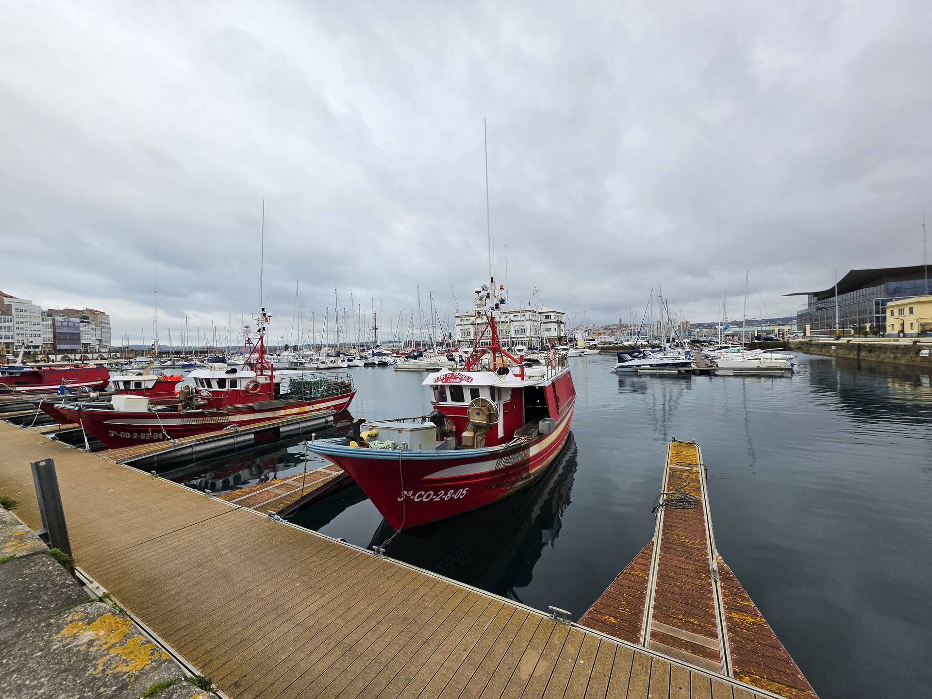 Barcos pesqueros rojos atracados en un puerto con cielo nublado y edificios al fondo, imagen marítima en la costa.