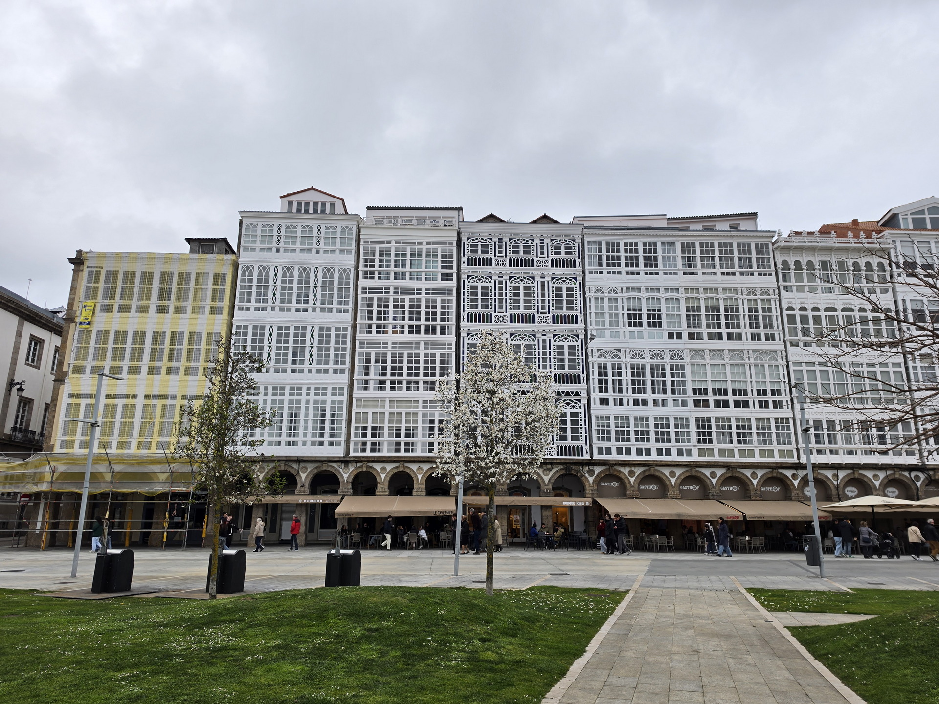 Edificios con galerías acristaladas en A Coruña, España, frente a una plaza con árboles y césped.