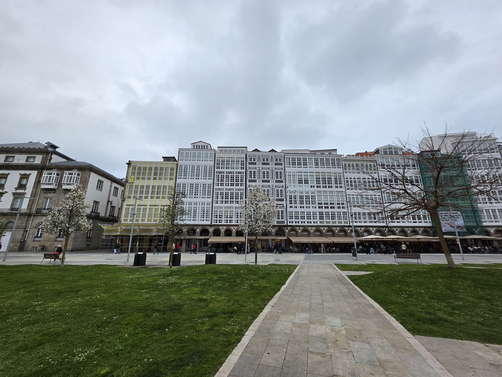 Edificios con galerías acristaladas en A Coruña, España, día nublado, césped y árboles sin hojas en primer plano.