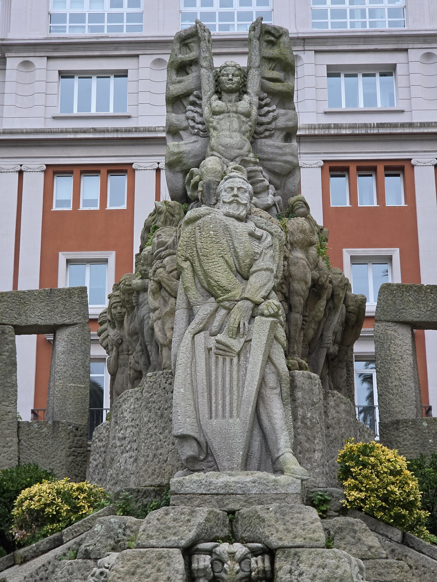Monumento de piedra en plaza de Lugo, A Coruña, con esculturas detalladas y edificio de fondo.