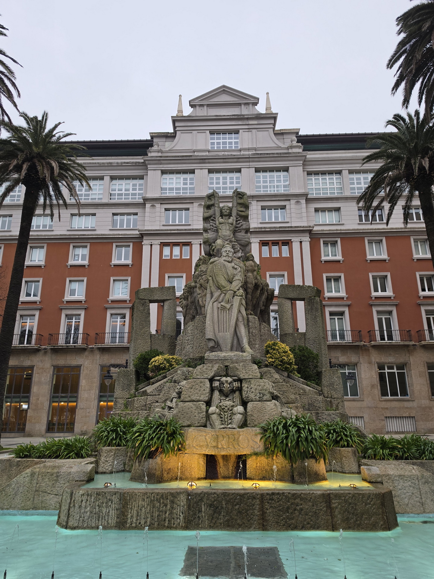 Fuente y escultura en plaza central, edificio clásico de fondo, rodeada de palmeras y jardines arbolados.