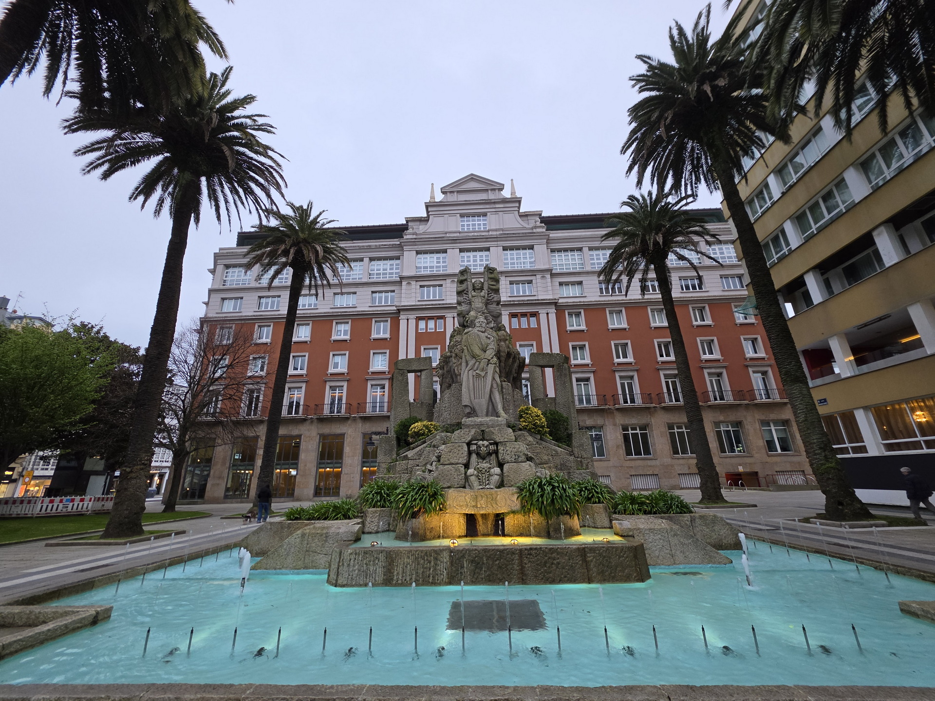 Fuente con esculturas y palmeras frente a un edificio clásico en una plaza urbana.