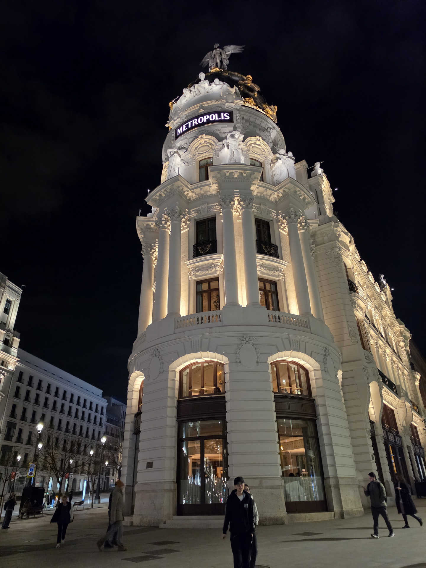 Edificio Metrópolis iluminado de noche en Madrid, con detalles arquitectónicos neoclásicos y estatua alada.
