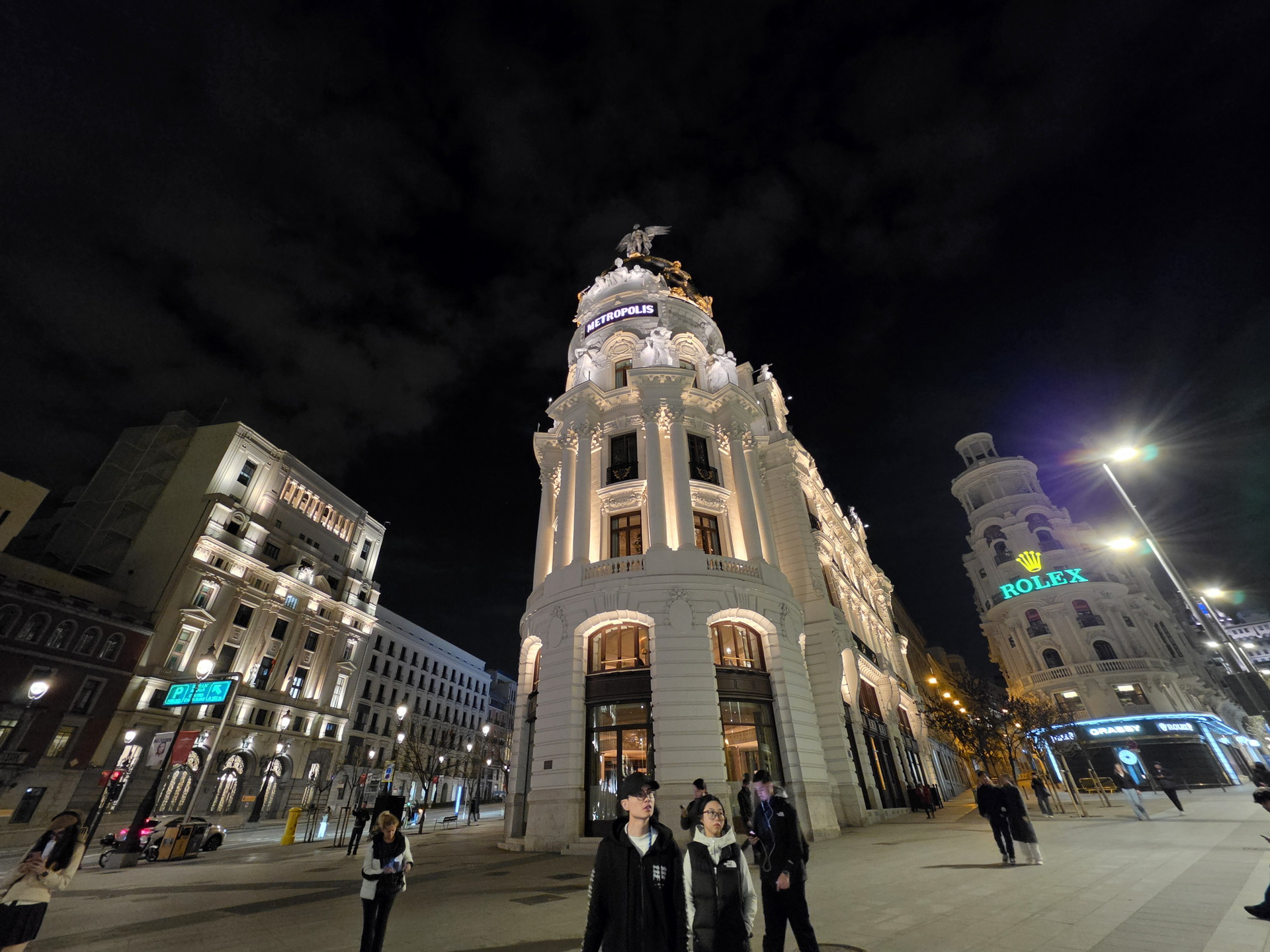 Edificio Metropolis iluminado de noche en Madrid, con personas en la calle y letreros visibles.