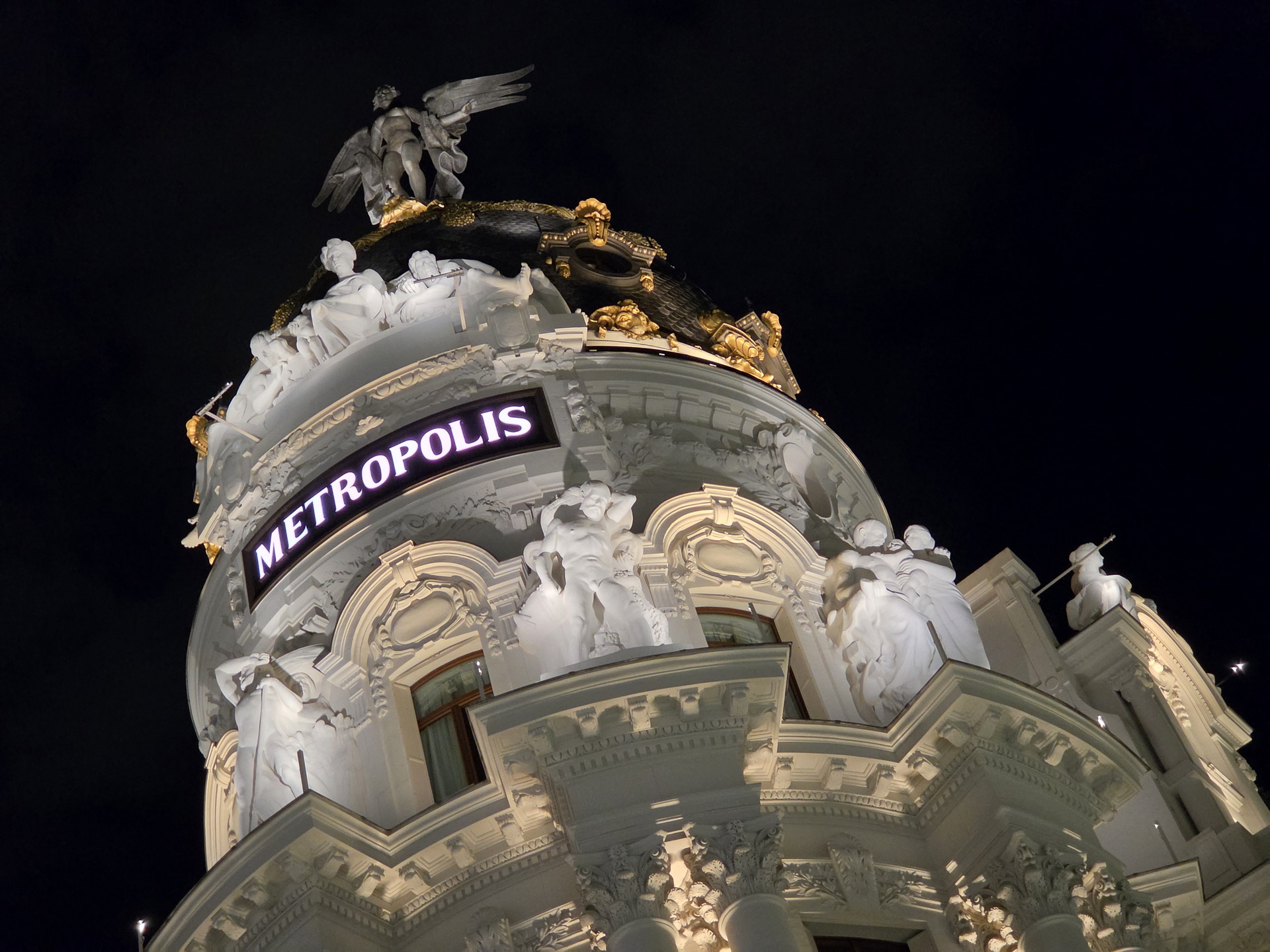 Edificio Metropolis iluminado de noche en Madrid, con detalle de esculturas clásicas y ángel alado en la cúspide.