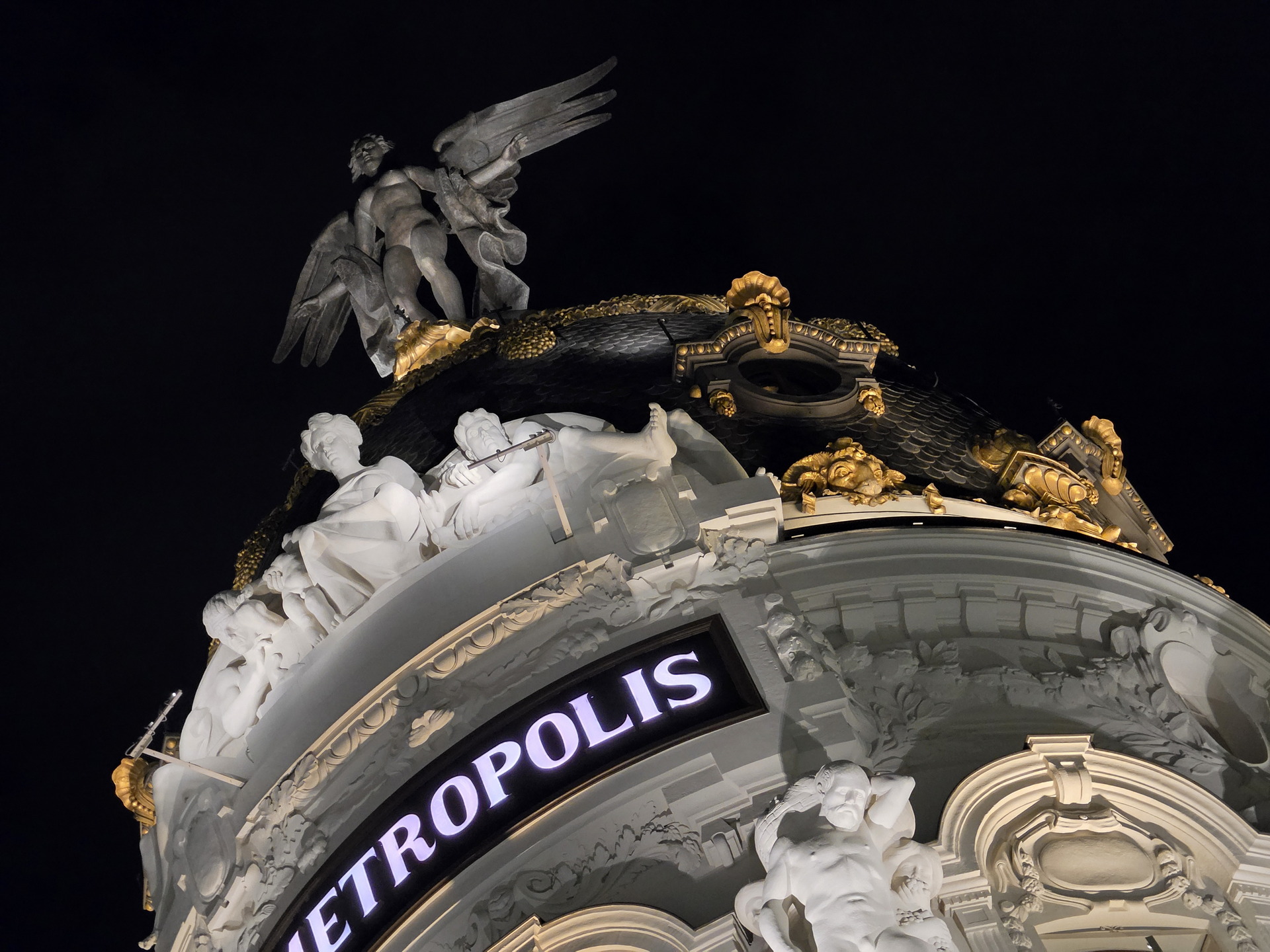Cúpula del edificio Metrópolis en Madrid iluminada de noche, con estatuas y detalles dorados.