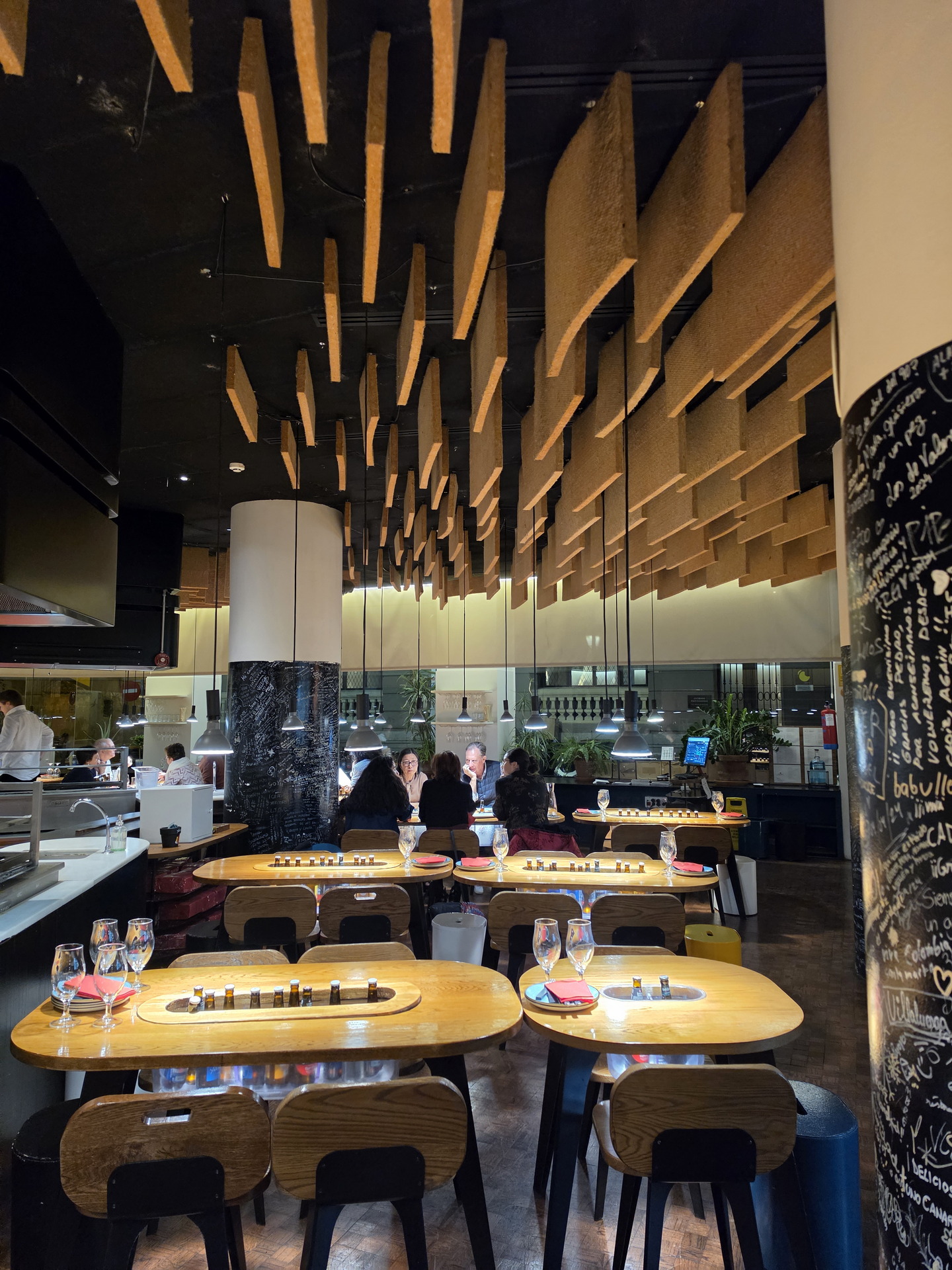 Restaurante moderno con mesas de madera, decoración colgante y ambiente acogedor. Comensales conversando.