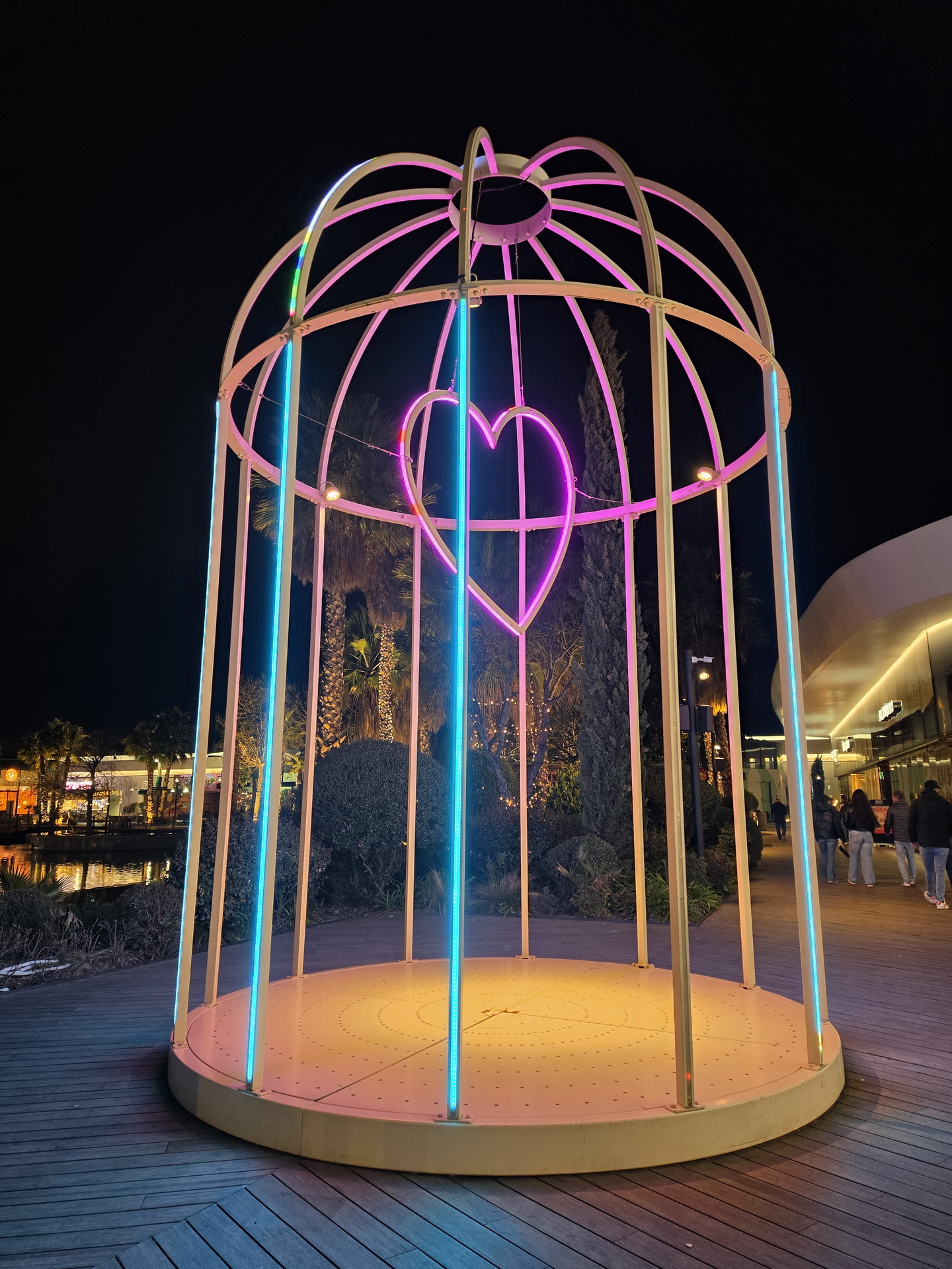 Jaula iluminada con neón y corazón rosa en su interior, ubicada en un paseo nocturno.