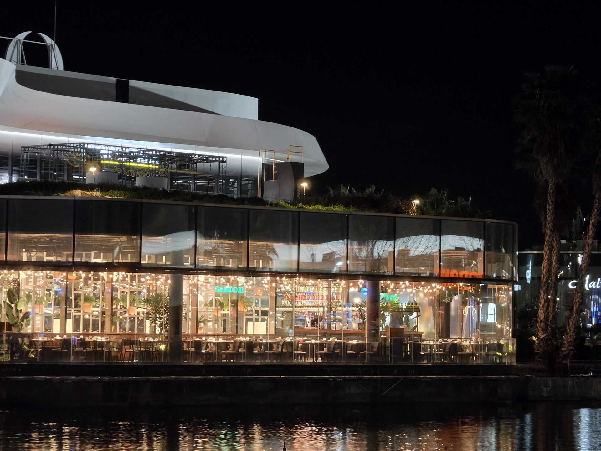 Restaurante moderno y luminoso frente al agua de noche, con decoración de luces y plantas en el interior.