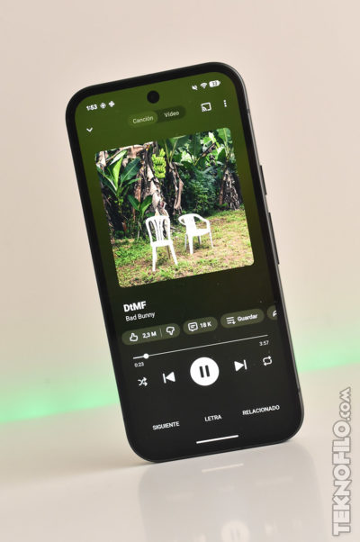 Teléfono mostrando canción DtMF de Bad Bunny en pantalla, con imagen de sillas blancas en jardín verde.