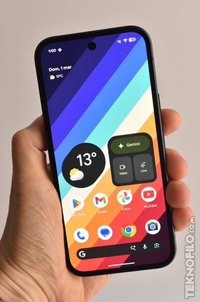 Teléfono móvil en mano con pantalla de inicio colorida mostrando widgets de clima y aplicaciones populares.
