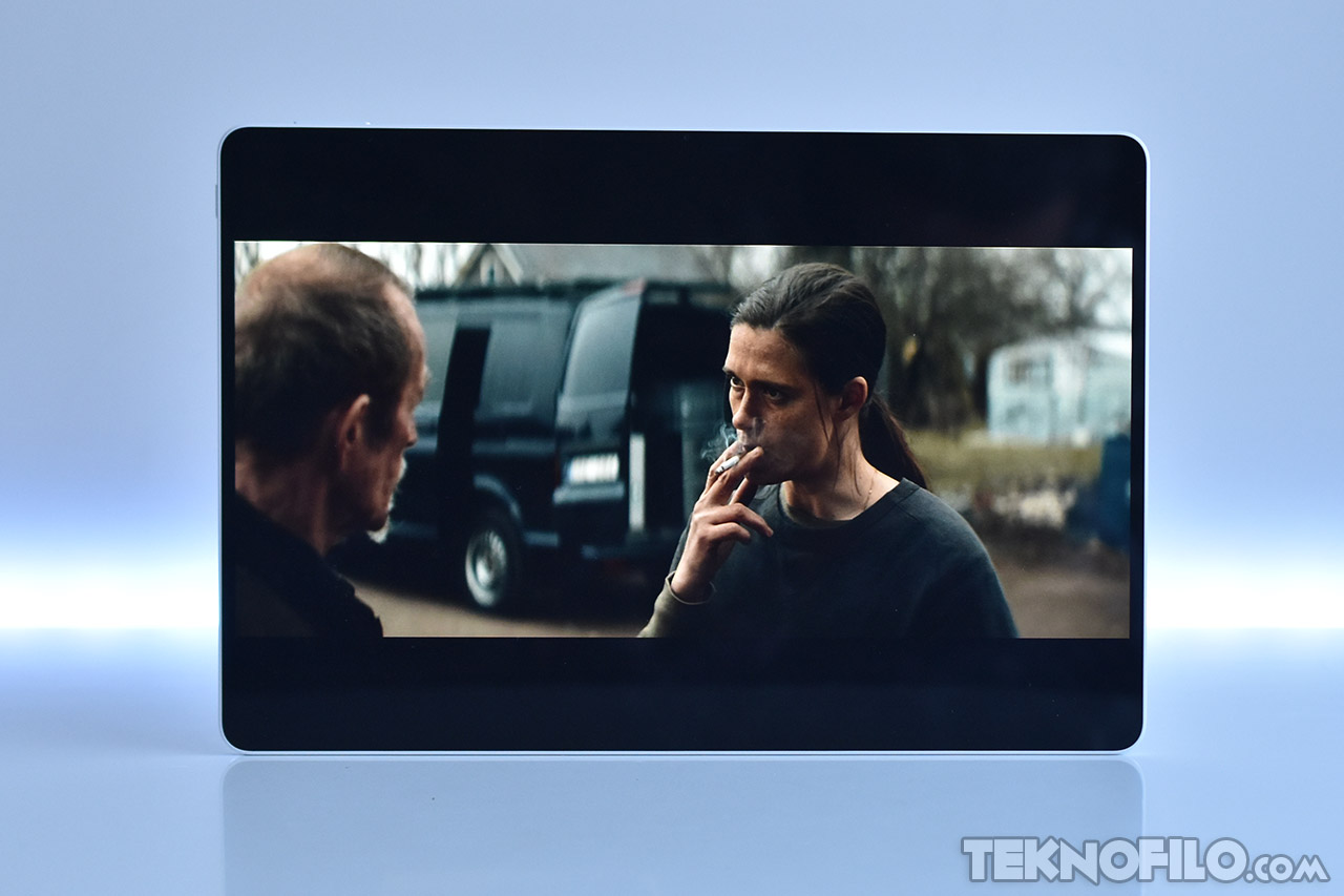 Pantalla de tablet mostrando una escena de película con dos personas conversando frente a una furgoneta negra.