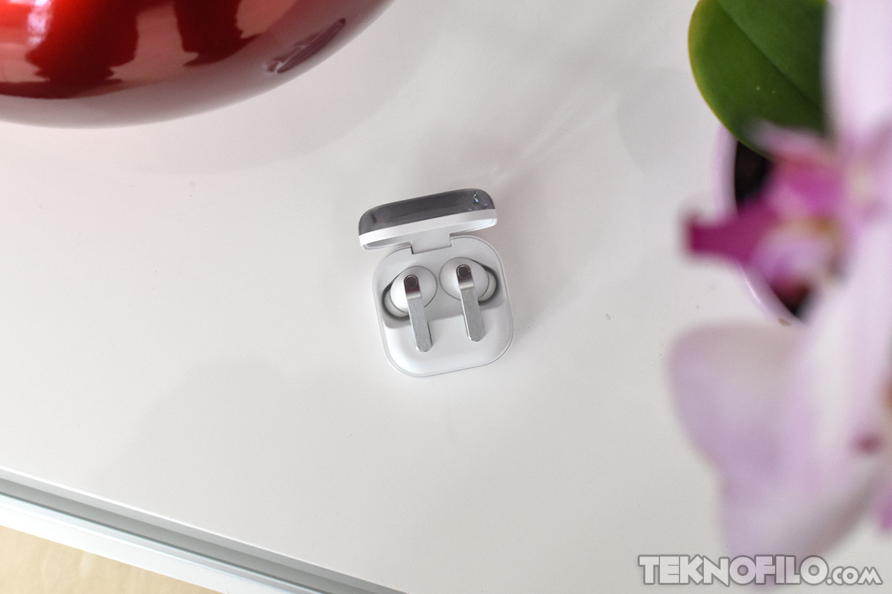 Auriculares inalámbricos en estuche sobre mesa blanca, junto a flor y objeto rojo. Ideal para música y llamadas.