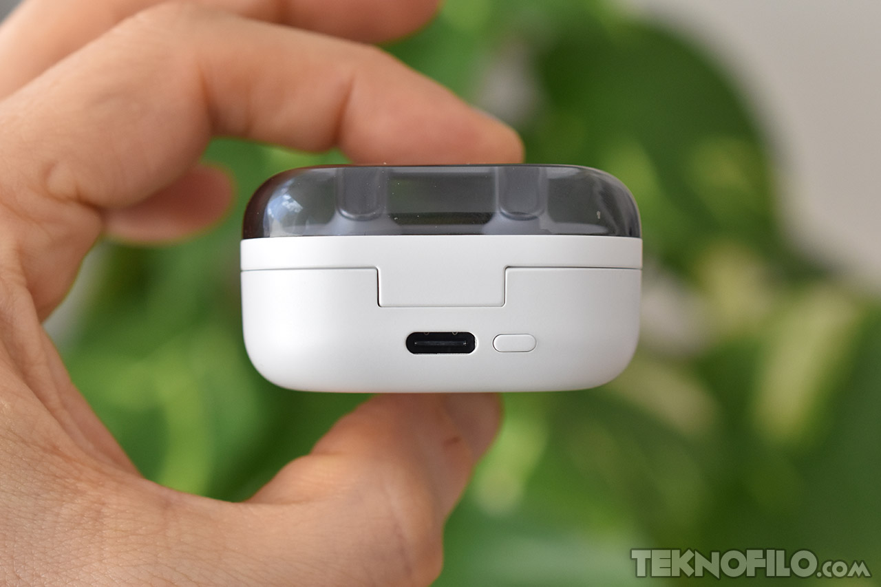 Estuche de carga de auriculares inalámbricos, blanco, sostenido por una mano, fondo de plantas verdes.