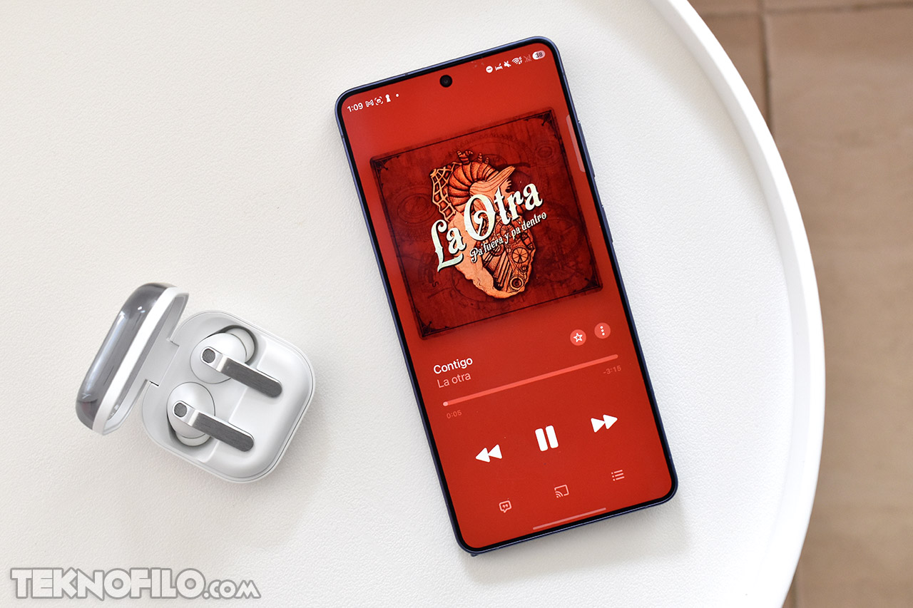Smartphone reproduciendo música Contigo de La Otra con audífonos inalámbricos en una mesa blanca.