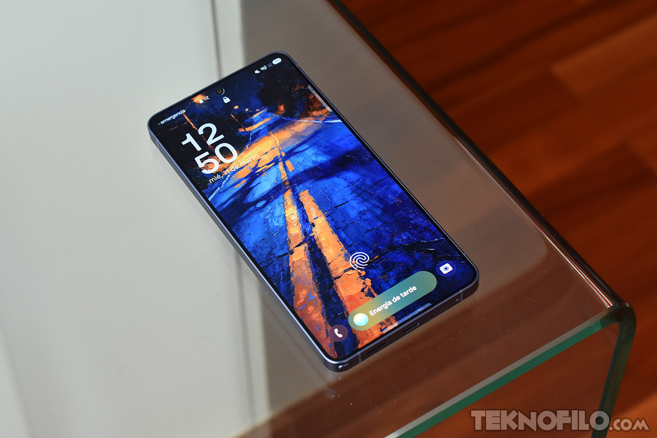 Smartphone sobre mesa de vidrio con pantalla encendida, mostrando un fondo artístico en tonos azul y naranja.